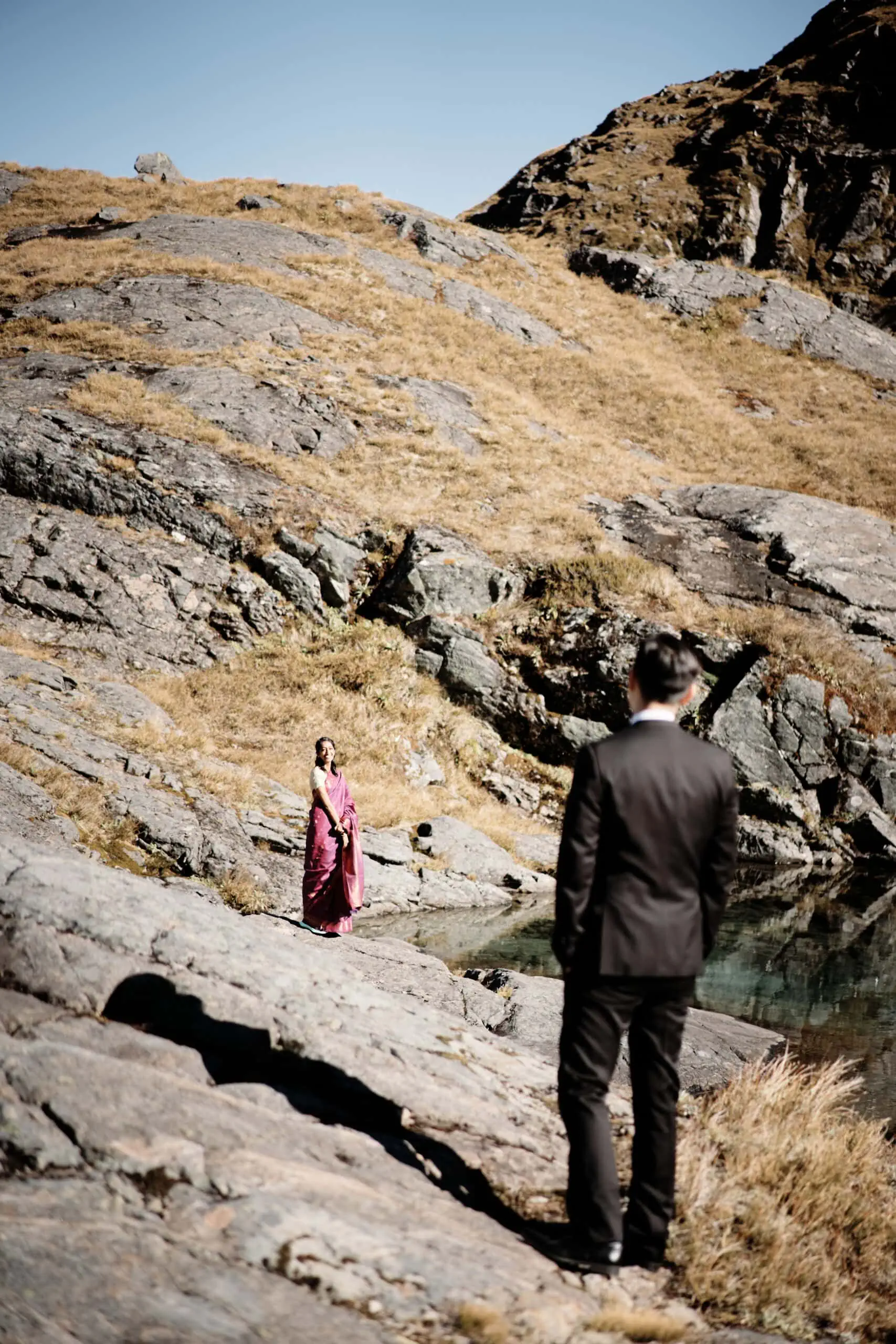 A man and woman standing next to a pond in the mountains.
