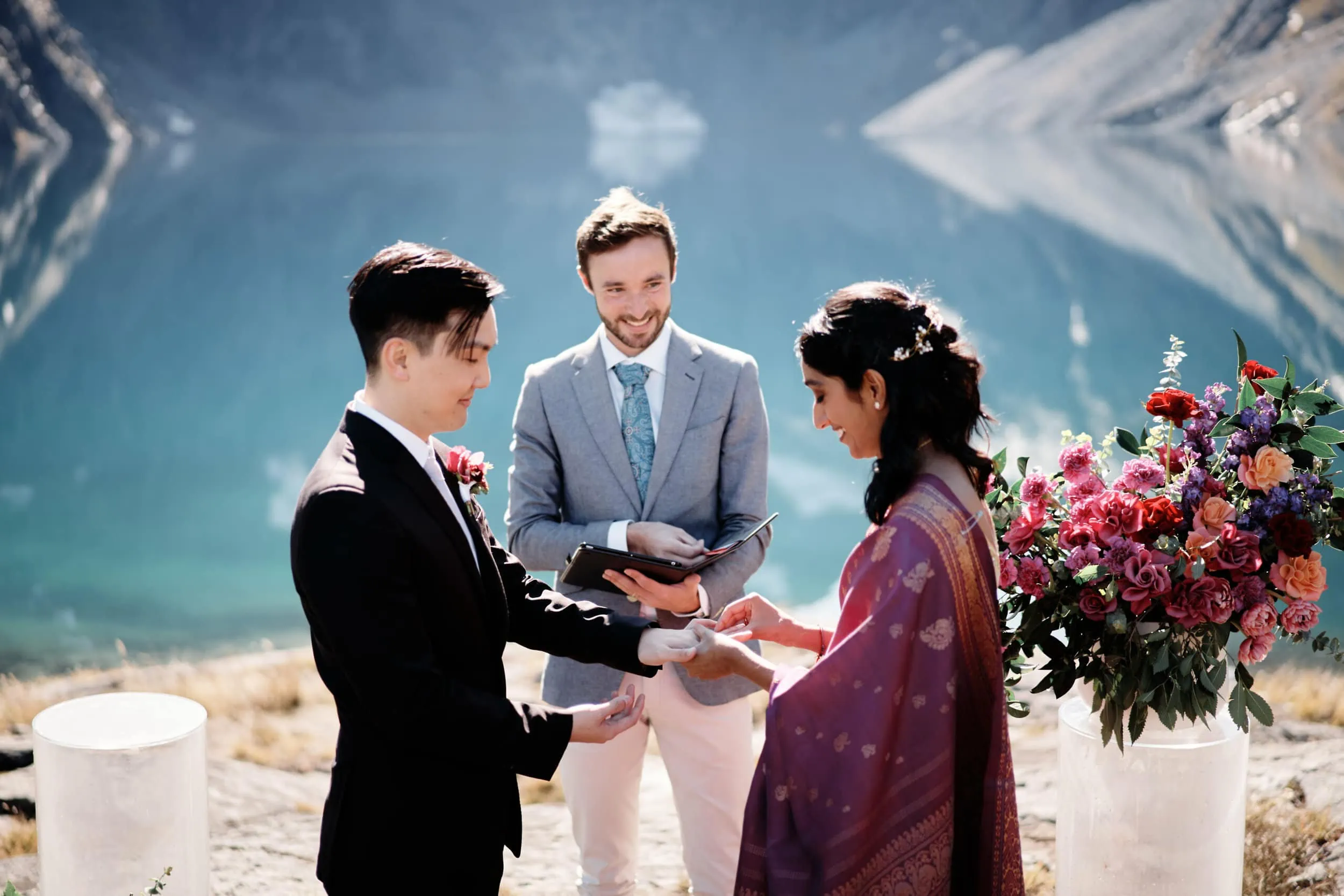 A bride and groom exchange rings in front of a lake.