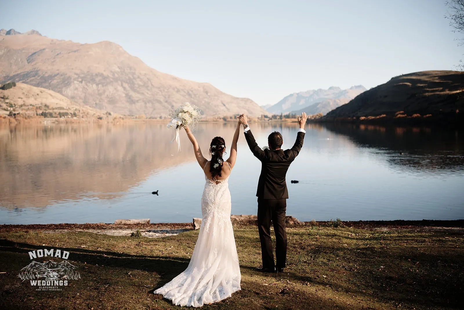 Connie and Andrew raising their hands in front of The Remarkables lake during their Heli Pre Wedding Shoot in New Zealand.