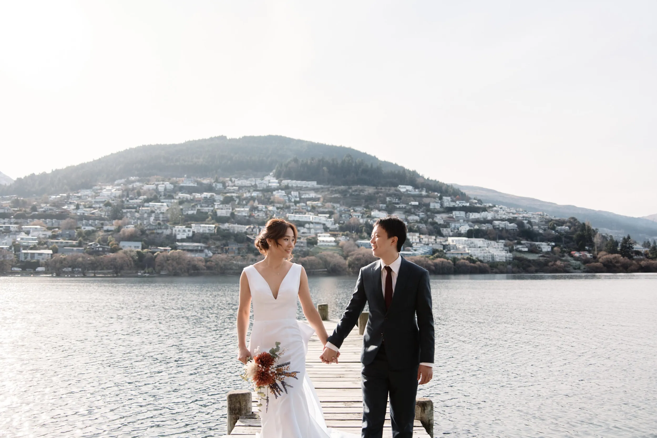 Yanzhi & Daphne's Queenstown Heli Pre Wedding Shoot captures a stunning moment of a bride and groom on a dock, with mountains in the background.