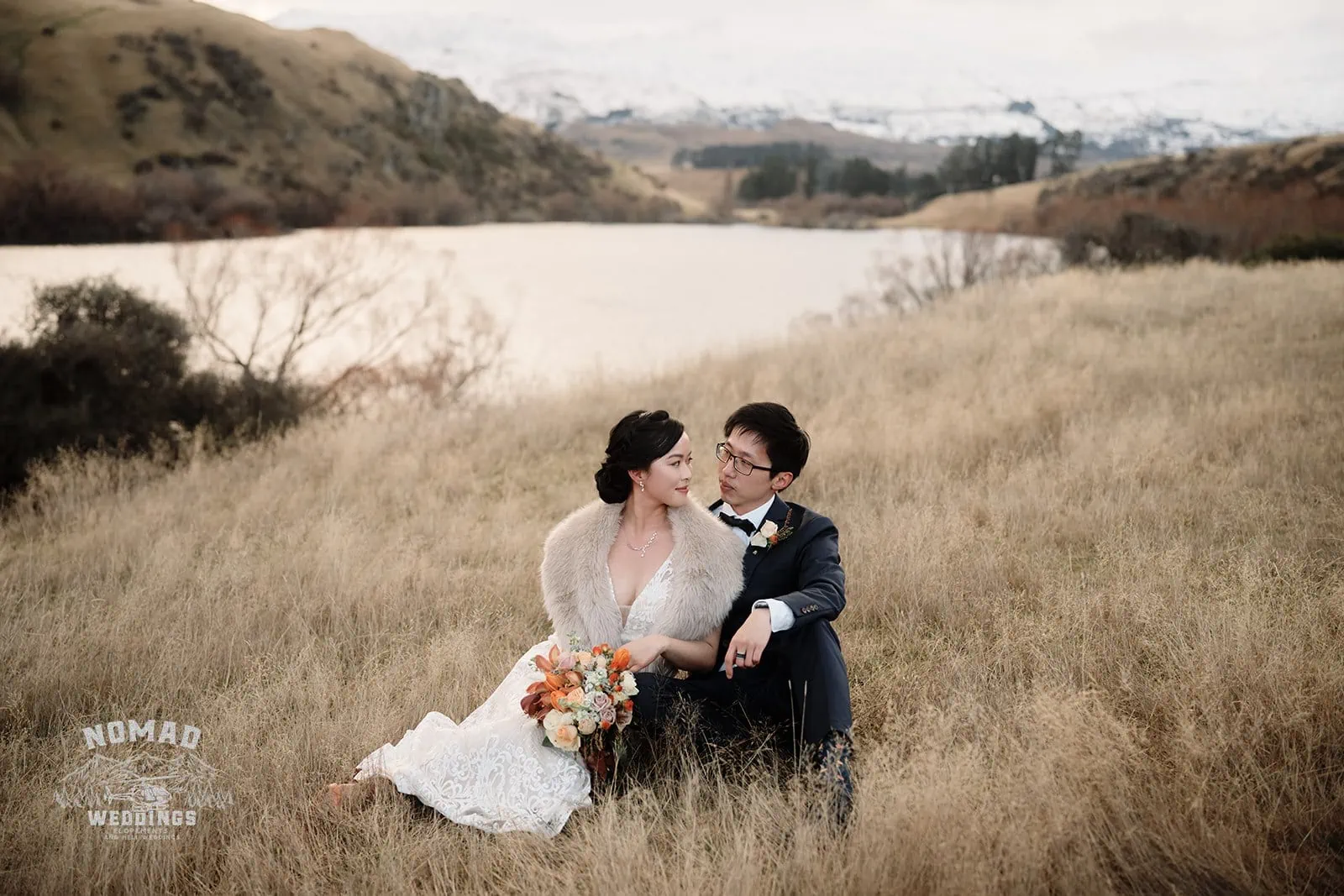Joanna and Tony's Pre-wedding Shoot in Queenstown, New Zealand, featuring a bride and groom sitting in a field near a lake.