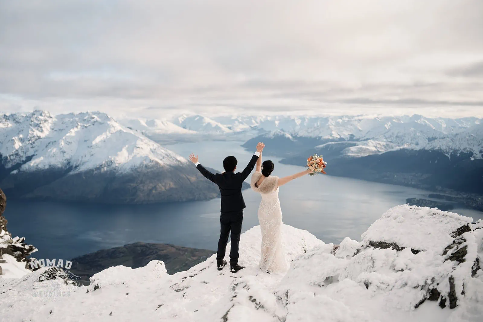 Joanna and Tony standing on top of a snowy mountain overlooking Lake Wanaka during their pre-wedding shoot in Queenstown, New Zealand.