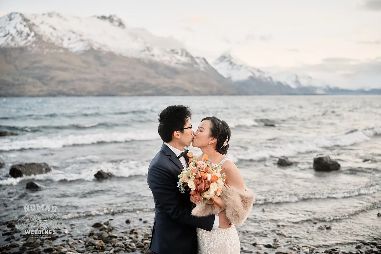 A pre-wedding shoot featuring Joanna and Tony, a bride and groom, sharing a kiss on the picturesque shore of Lake Wanaka in Queenstown, New Zealand.