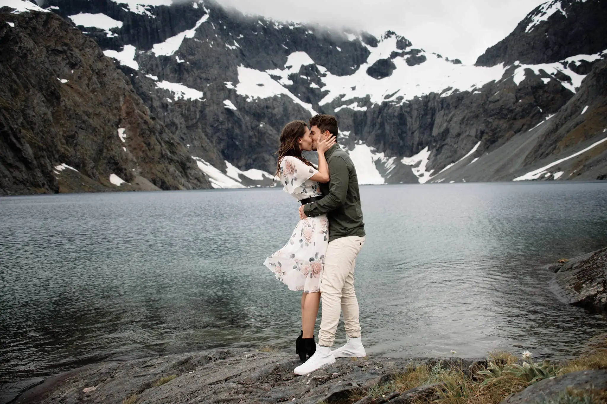 Scott and Hayley kiss in front of a lake with mountains in the background.