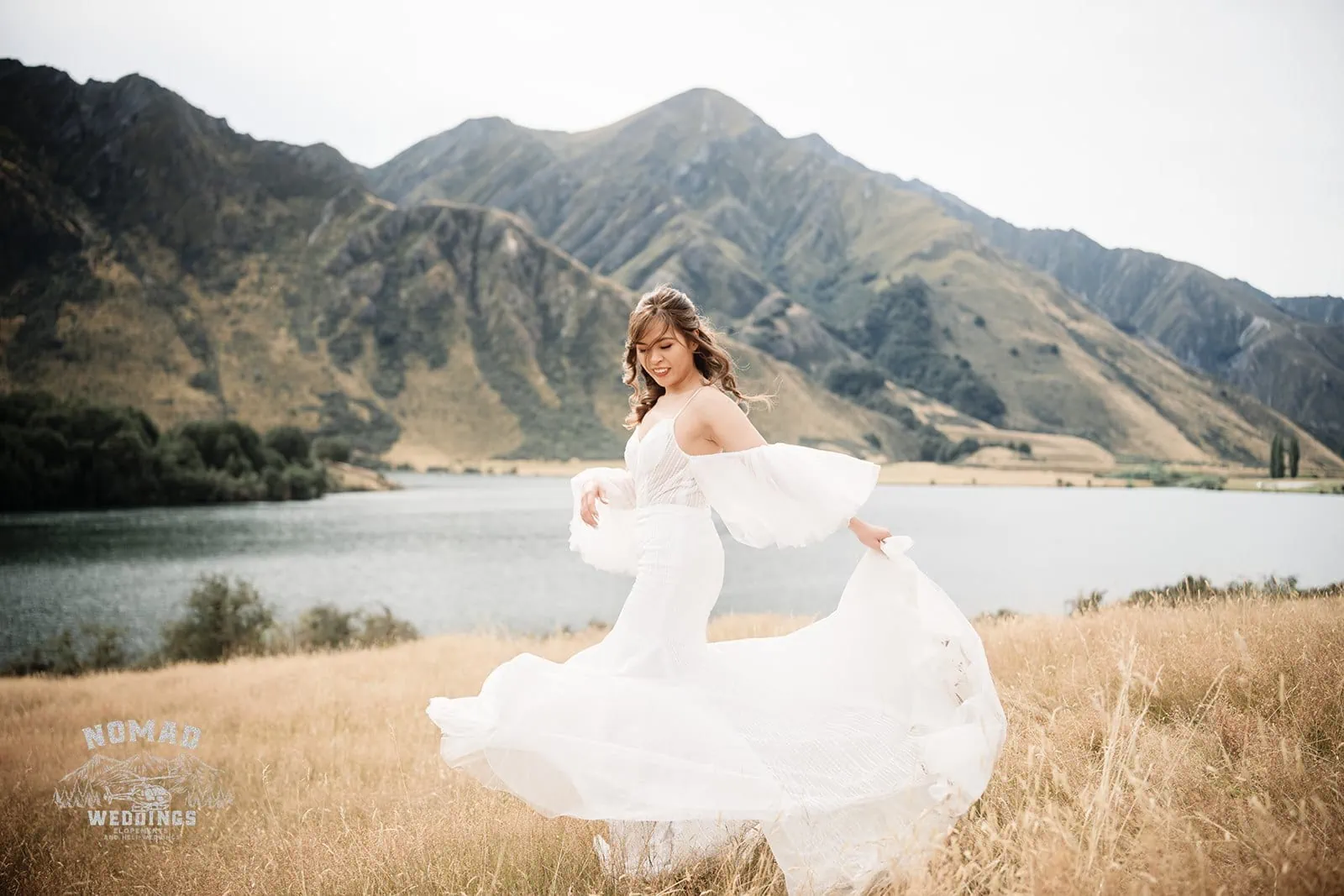 A bride in a white dress is standing near a lake during Nhi & Nicholas' Queenstown NZ Cecil Peak Pre-Wedding Shoot.