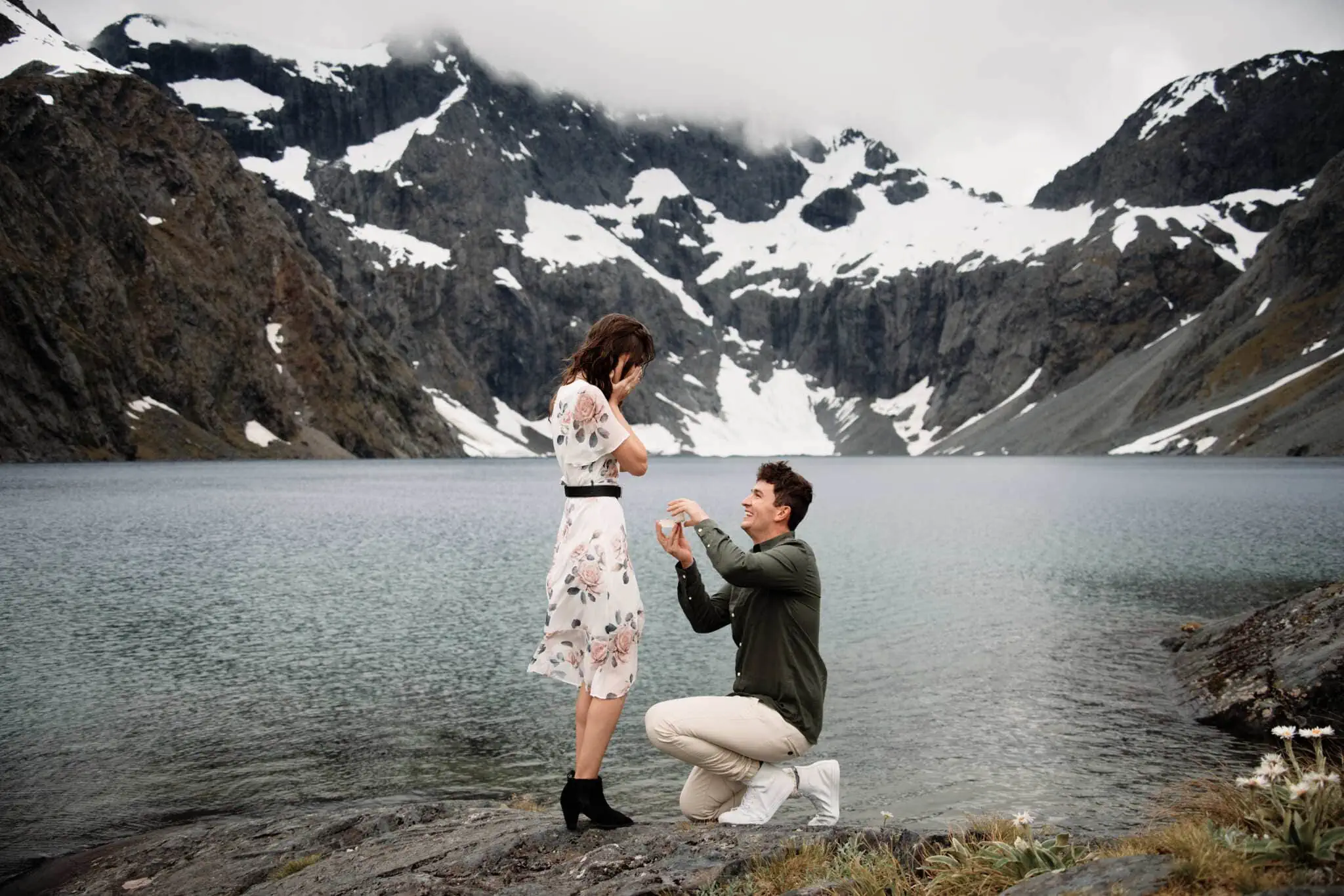 Scott and Hayley are a couple posing by a lake with mountains as their backdrop.