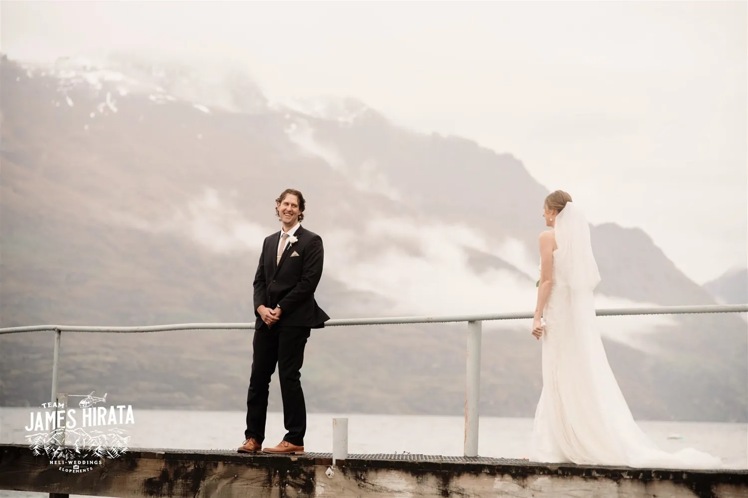 Hannah and Ross Elopement Wedding in Queenstown New Zealand: A bride and groom standing on a dock in front of mountains.