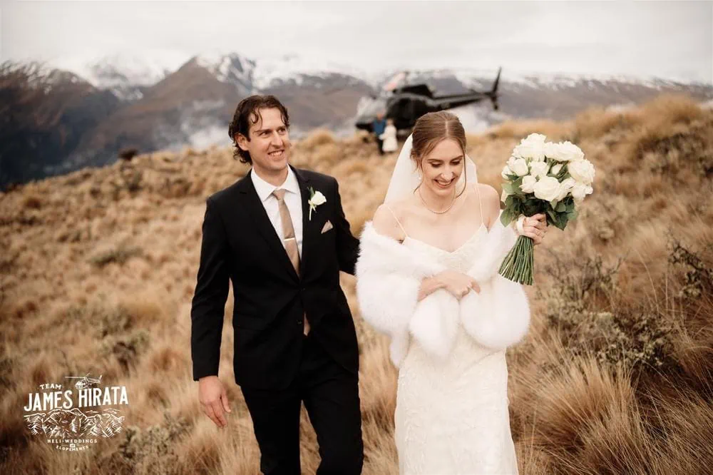 Hannah and Ross's elopement wedding in Queenstown, New Zealand, features a bride and groom walking through a grassy field with mountains in the background.