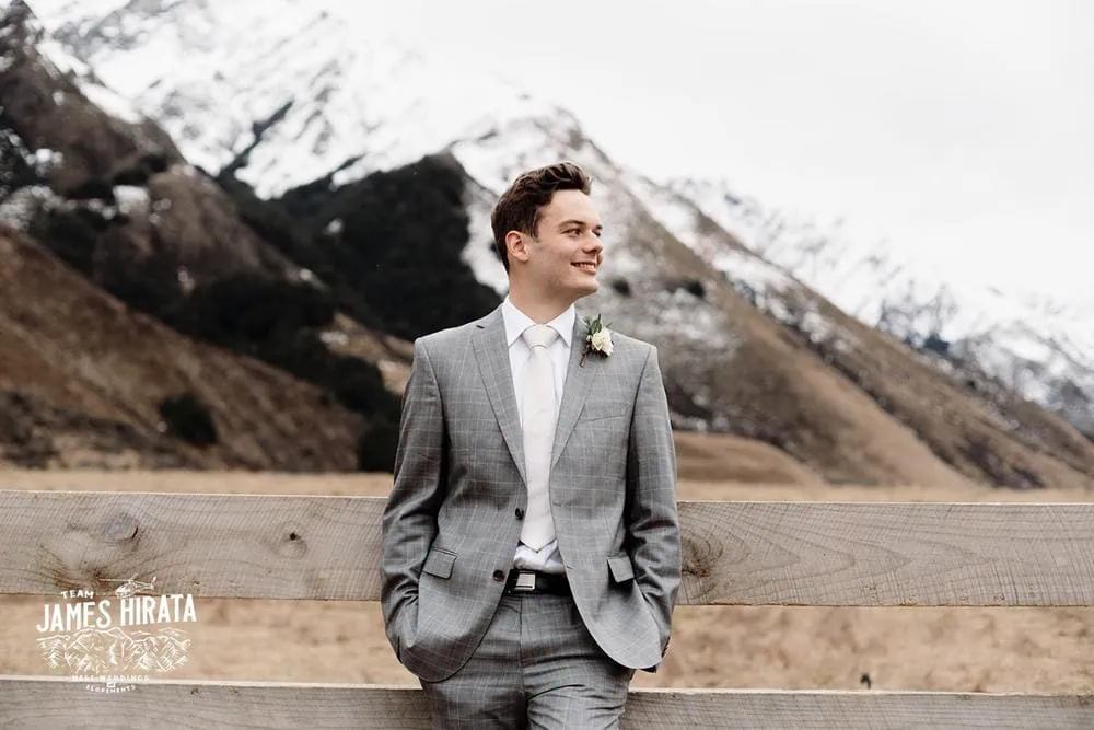 Regan & Jake's Queenstown elopement wedding featuring a groom in a suit leaning against a fence, with mountains in the background.