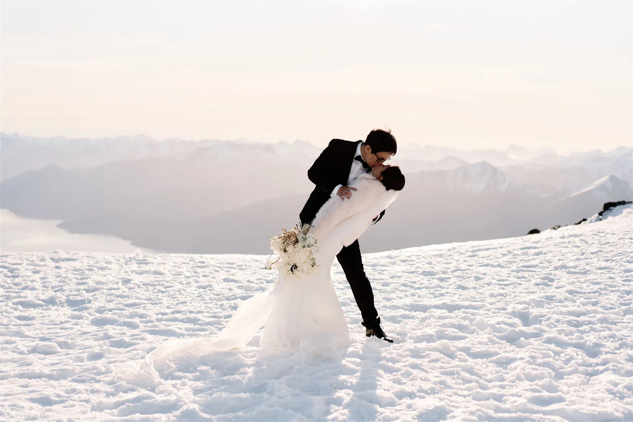 Queenstown Elopement Heli Wedding Photographer クイーンズタウン結婚式 | A bride and groom kiss on top of a snow covered mountain in Queenstown.