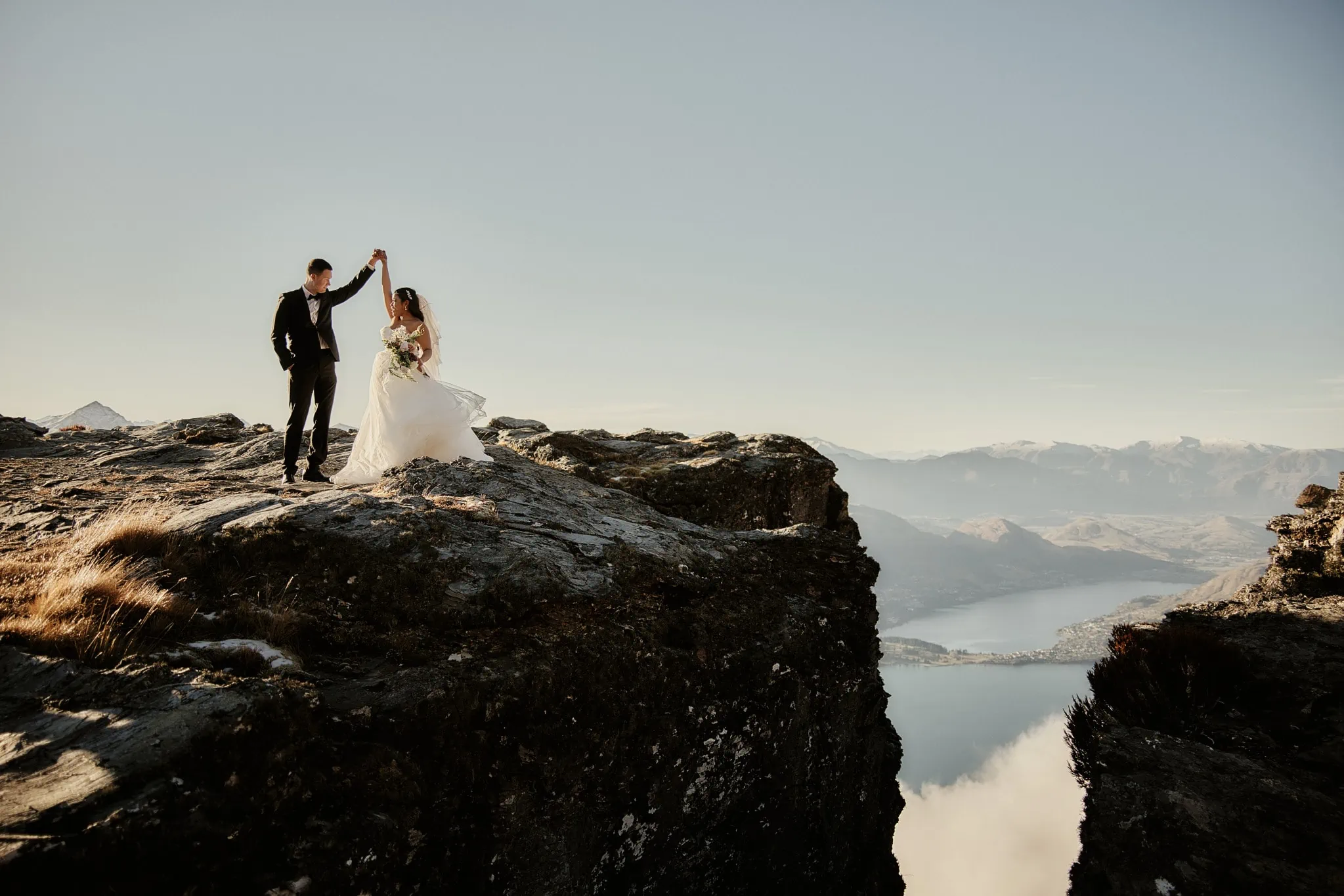 Queenstown New Zealand Elopement Wedding Photographer - Amy and Callum's Queenstown Heli Pre Wedding Shoot captures a bride and groom standing on top of a cliff overlooking Lake Wanaka.