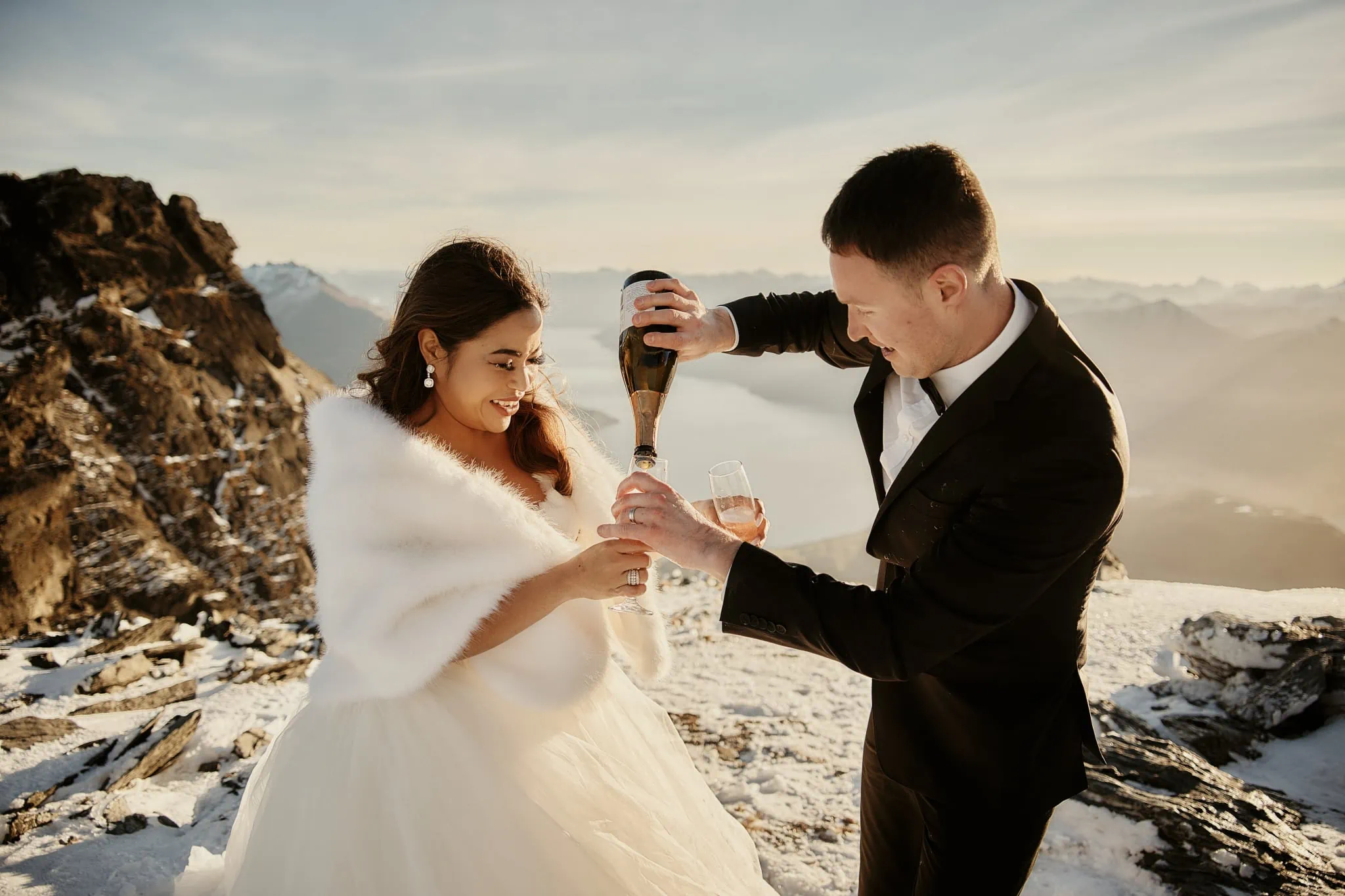 Queenstown New Zealand Elopement Wedding Photographer - Amy and Callum's Queenstown Heli Pre Wedding Shoot captures the couple pouring champagne on top of a snowy mountain.