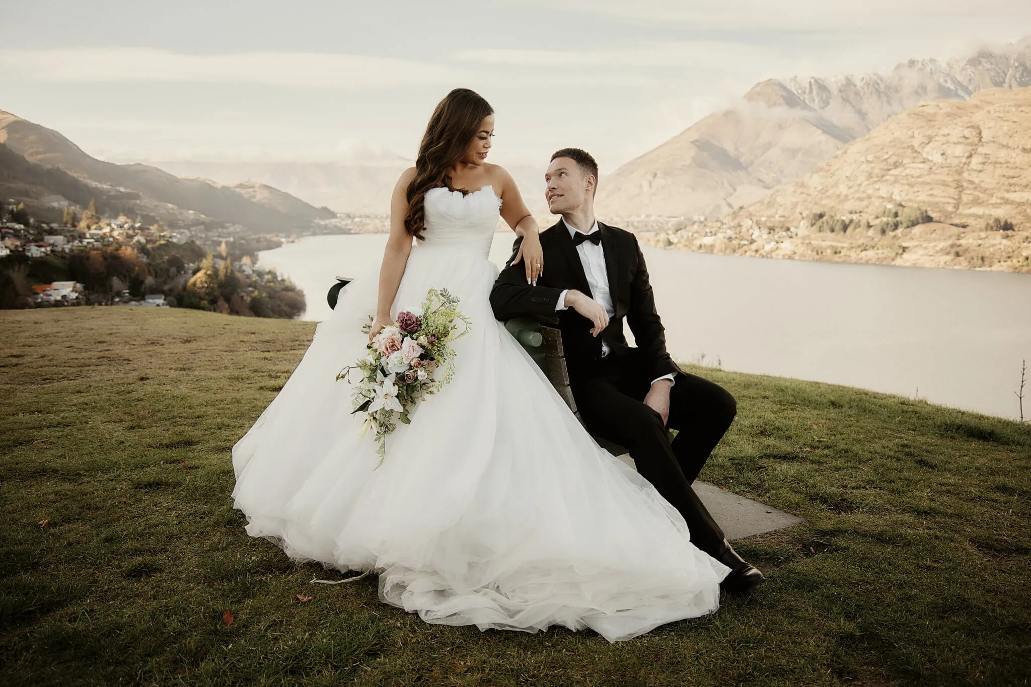 Queenstown New Zealand Elopement Wedding Photographer - Amy and Callum's Queenstown Heli Pre Wedding Shoot features a bride and groom sitting on a bench in front of a lake.