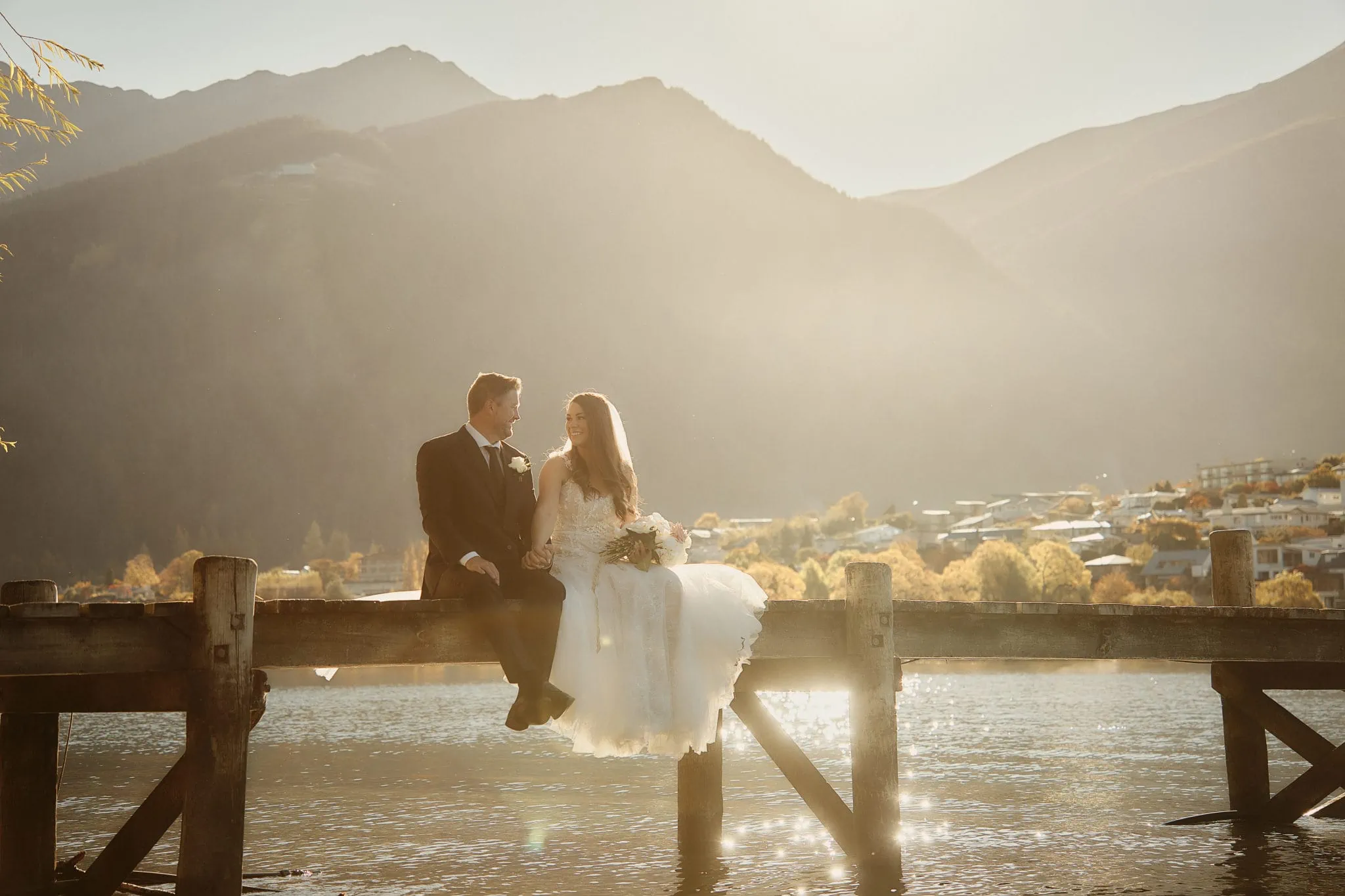 Queenstown New Zealand Elopement Wedding Photographer - Ashleigh and Anthony's Roy's Peak Heli Elopement Wedding, bride and groom sitting on a pier in front of a lake.