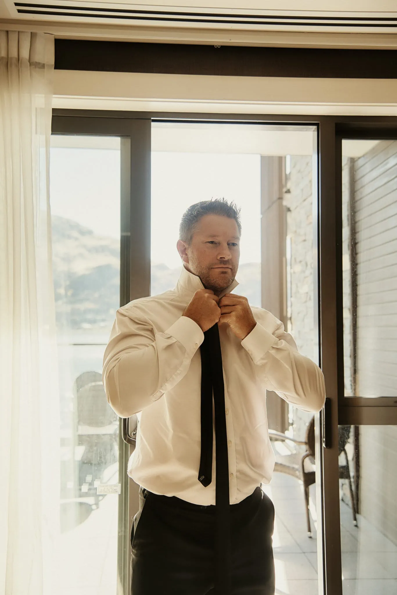 Queenstown New Zealand Elopement Wedding Photographer - A man adjusting his tie in front of a window during Ashleigh and Anthony's Roy's Peak Heli Elopement Wedding.