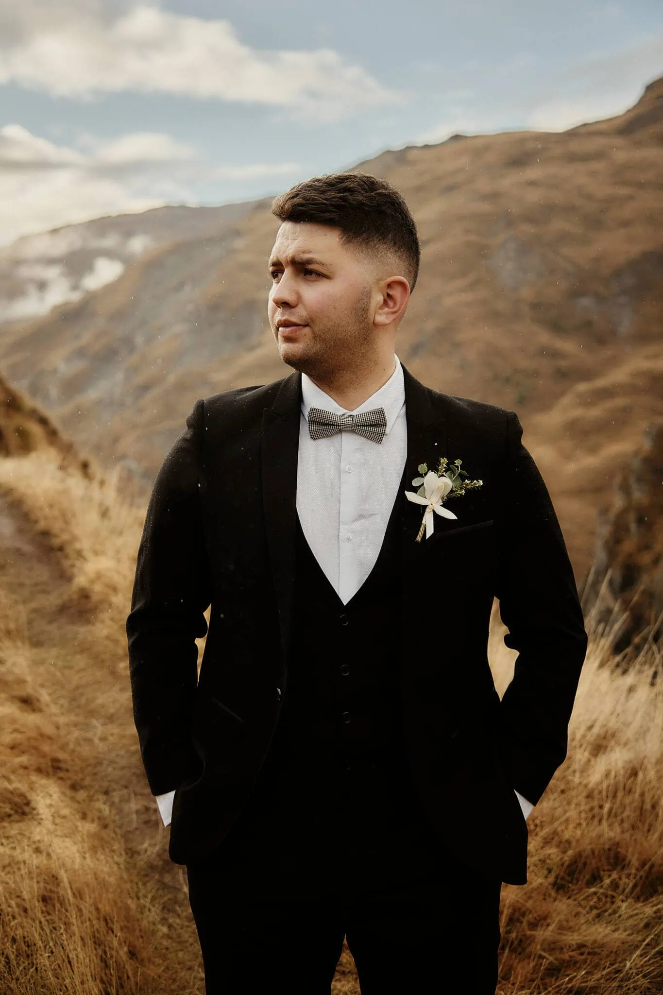 Queenstown New Zealand Elopement Wedding Photographer - Caitiann and Kiyo's elopement wedding at Coronet Peak features a groom in a black suit standing in a grassy field.