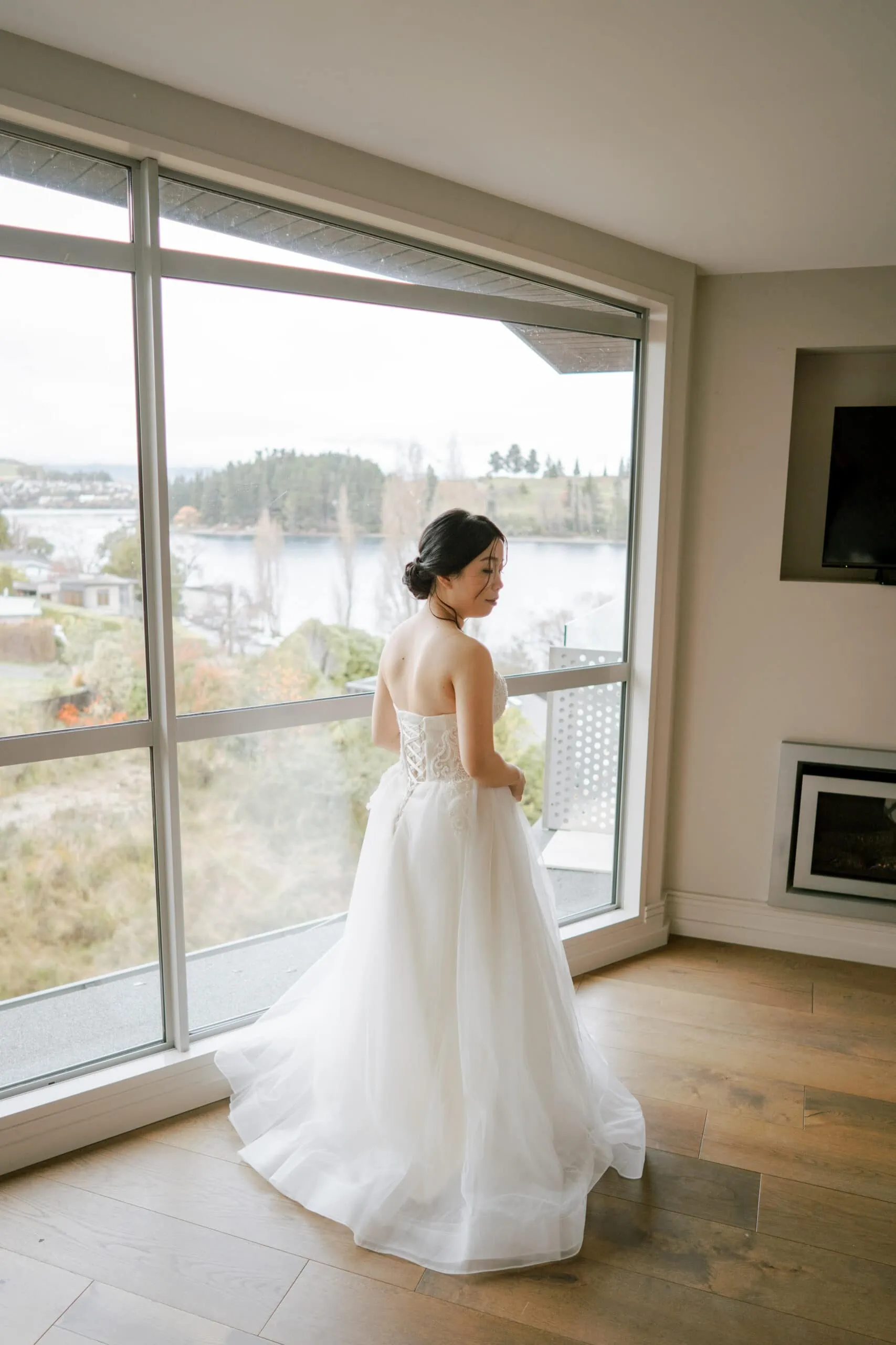 Queenstown New Zealand Elopement Wedding Photographer - A bride in a wedding dress standing in front of a window at Lam and Wendy's Kamana Lakehouse Wedding.
