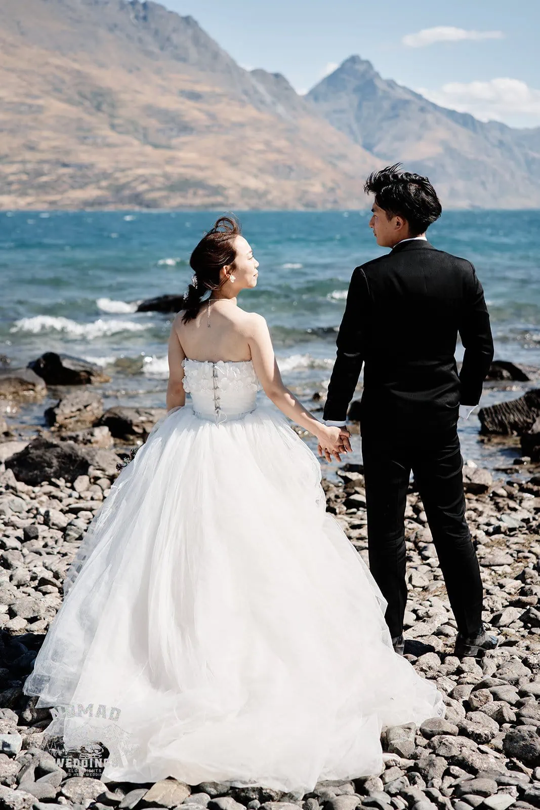 Queenstown New Zealand Elopement Wedding Photographer - A bride and groom are holding hands on a rocky beach in New Zealand for the Summer Edition - Ground Guide to the Seasons in Queenstown.