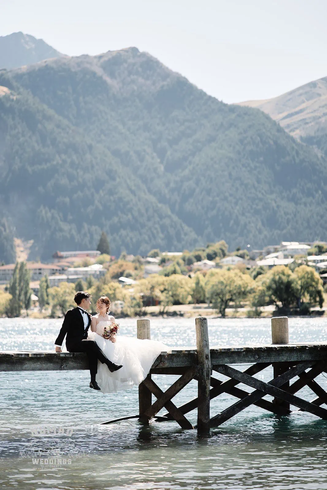Queenstown New Zealand Elopement Wedding Photographer - A bride and groom sitting on a dock with mountains in the background, showcasing the summer season in Queenstown.