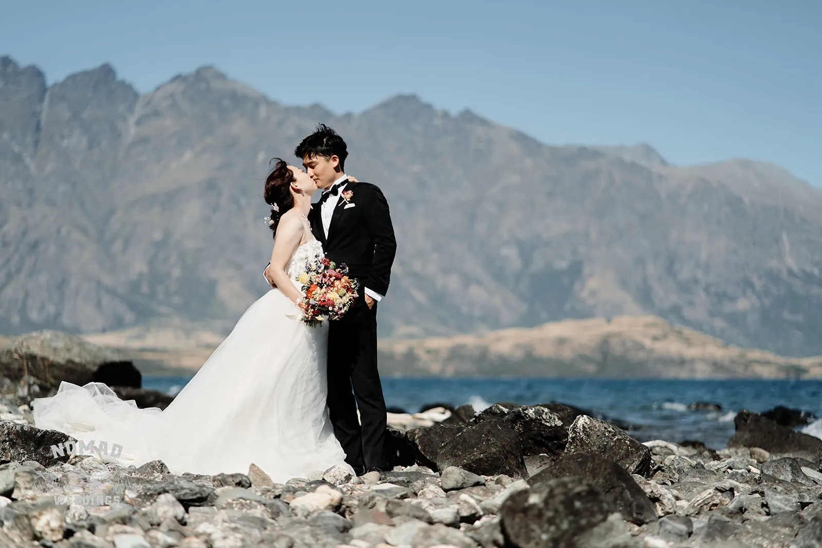 Queenstown New Zealand Elopement Wedding Photographer - A bride and groom standing on a rocky beach with mountains in the background - QUEENSTOWN SEASONS.