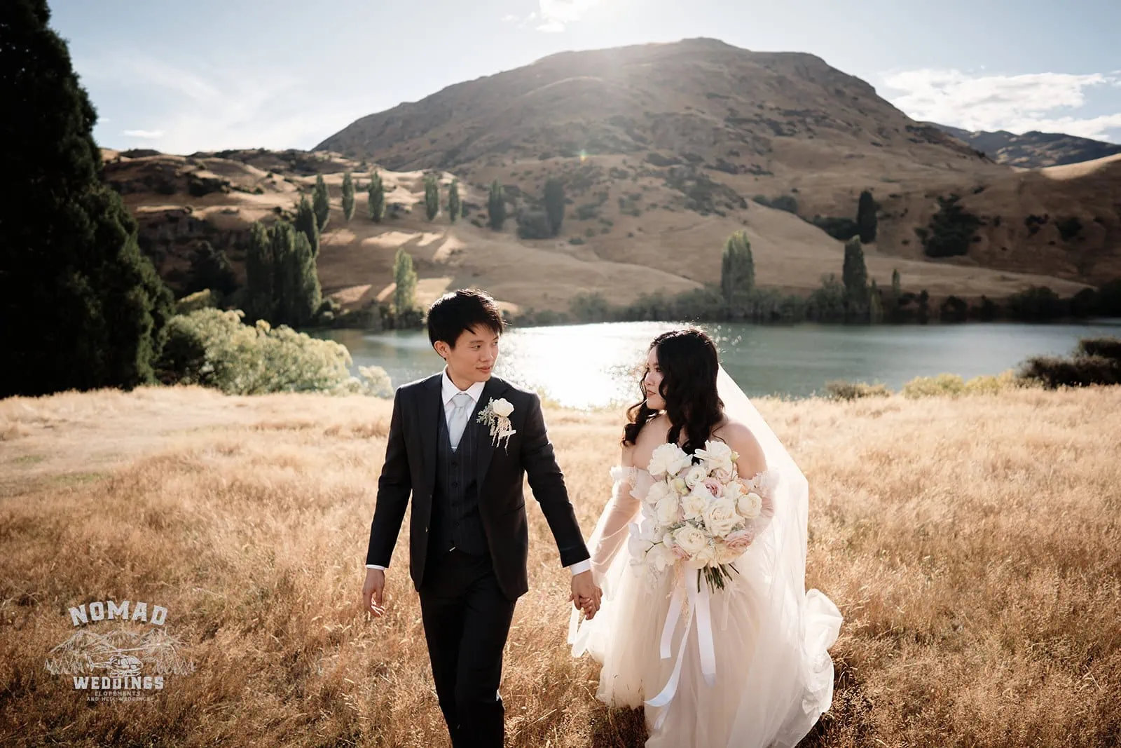 Queenstown New Zealand Elopement Wedding Photographer - A couple strolling through a scenic field by a lake in Queenstown during the summer.