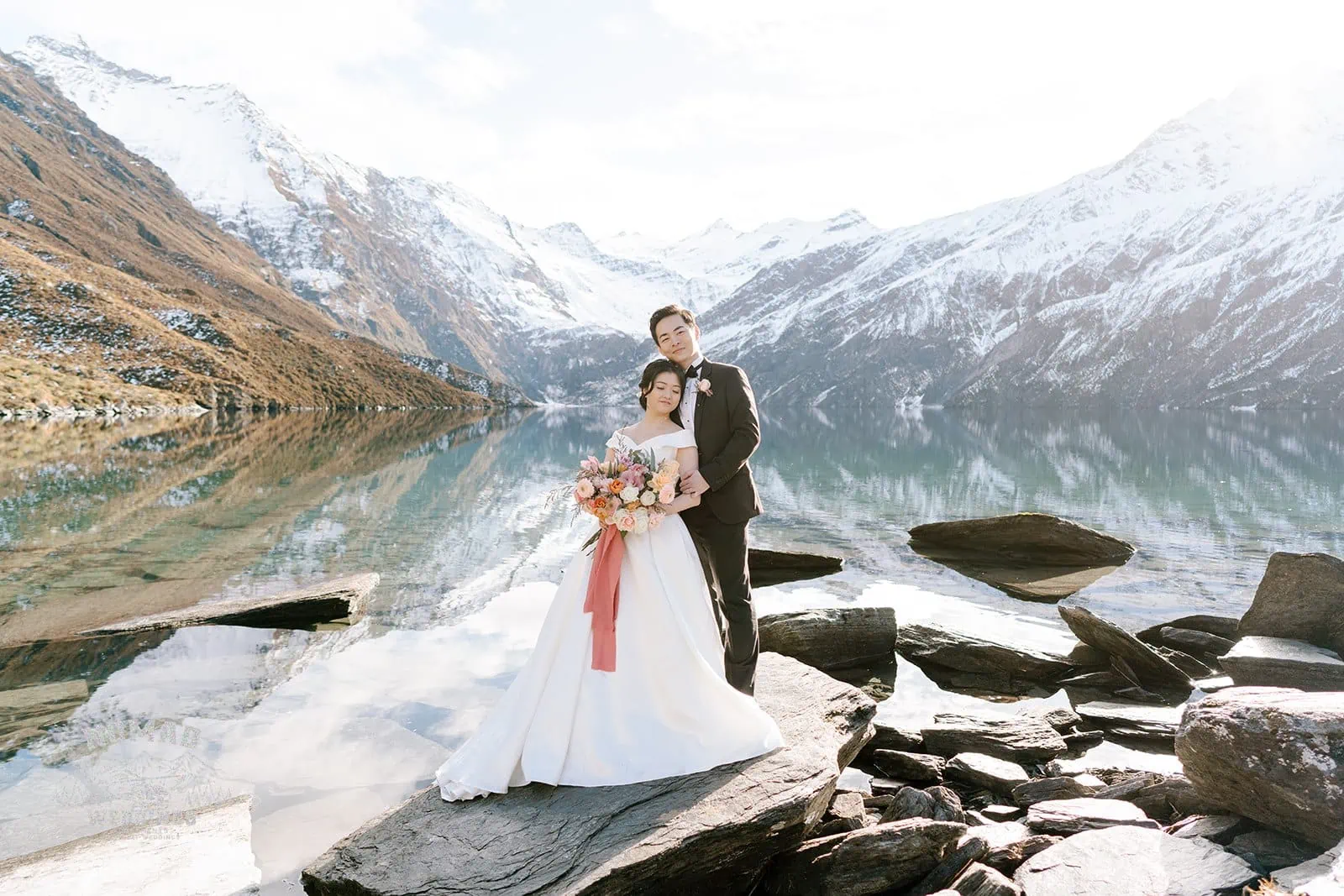 Queenstown New Zealand Elopement Wedding Photographer - Bo and Junyi, a bride and groom, having a Heli Pre Wedding Shoot with 4 Landings in New Zealand, standing on rocks in front of a lake.