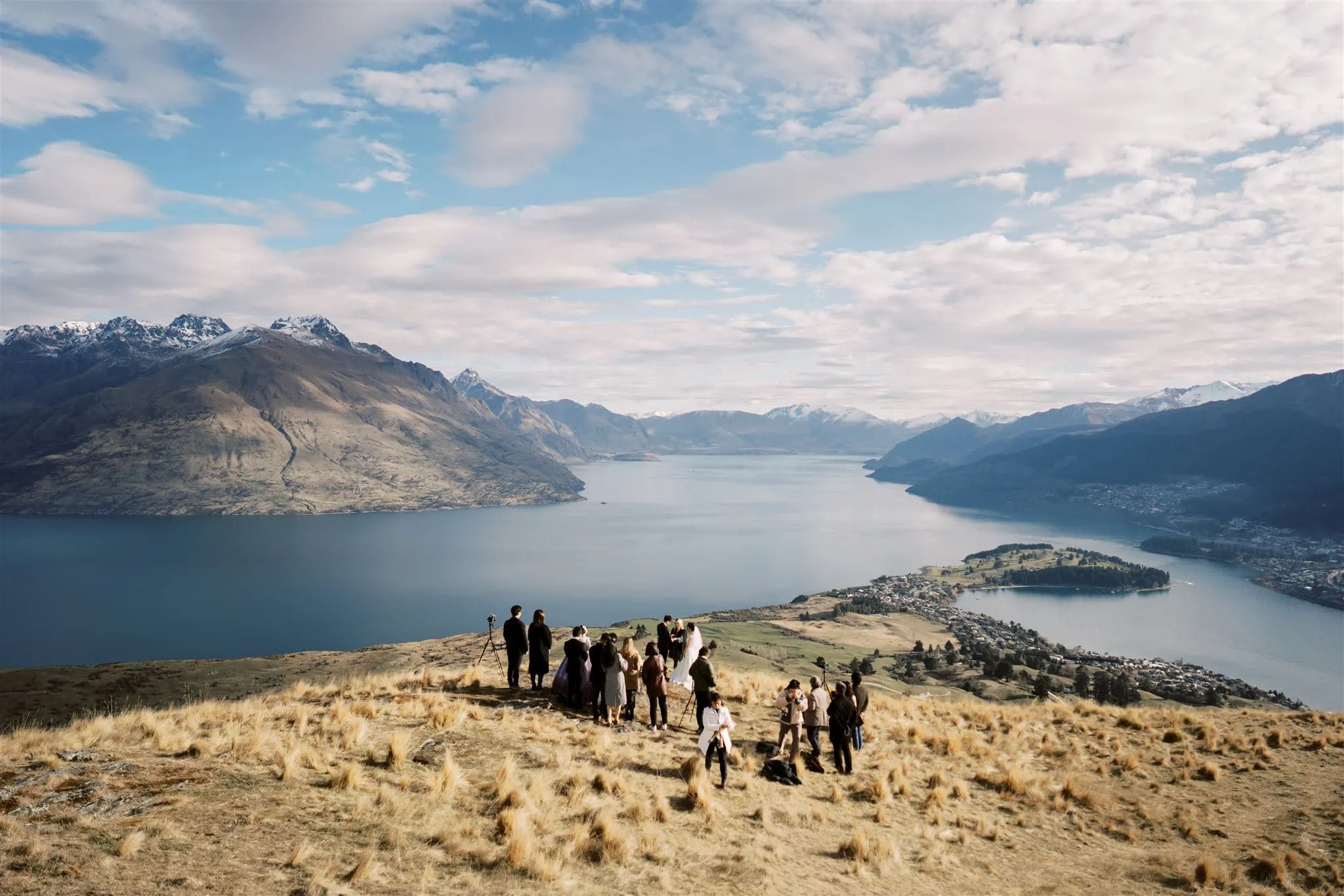 Queenstown New Zealand Elopement Wedding Photographer - A group of people standing on top of a hill overlooking Lake Wakatipu, comparing the costs of elopement and traditional weddings.