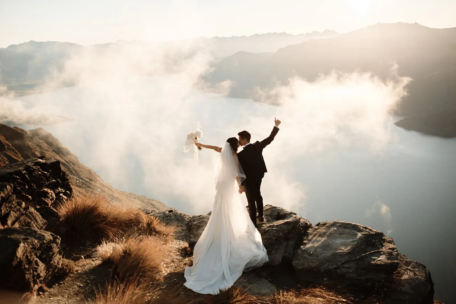 Queenstown New Zealand Elopement Wedding Photographer - The Ultimate Queenstown Elopement Wedding Guide featuring a bride and groom atop a mountain, overlooking the clouds and lake at sunset