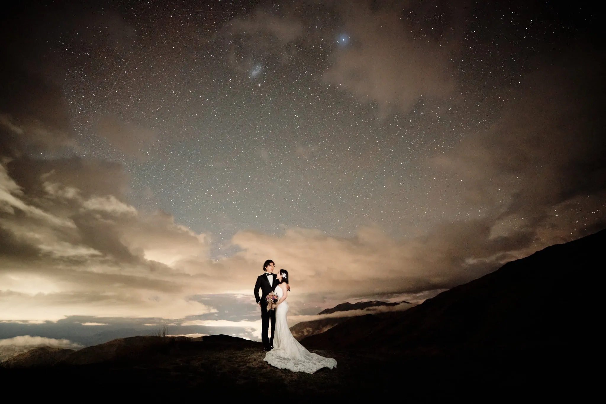 Queenstown New Zealand Elopement Wedding Photographer - A bride and groom standing on top of a mountain under a starry night.