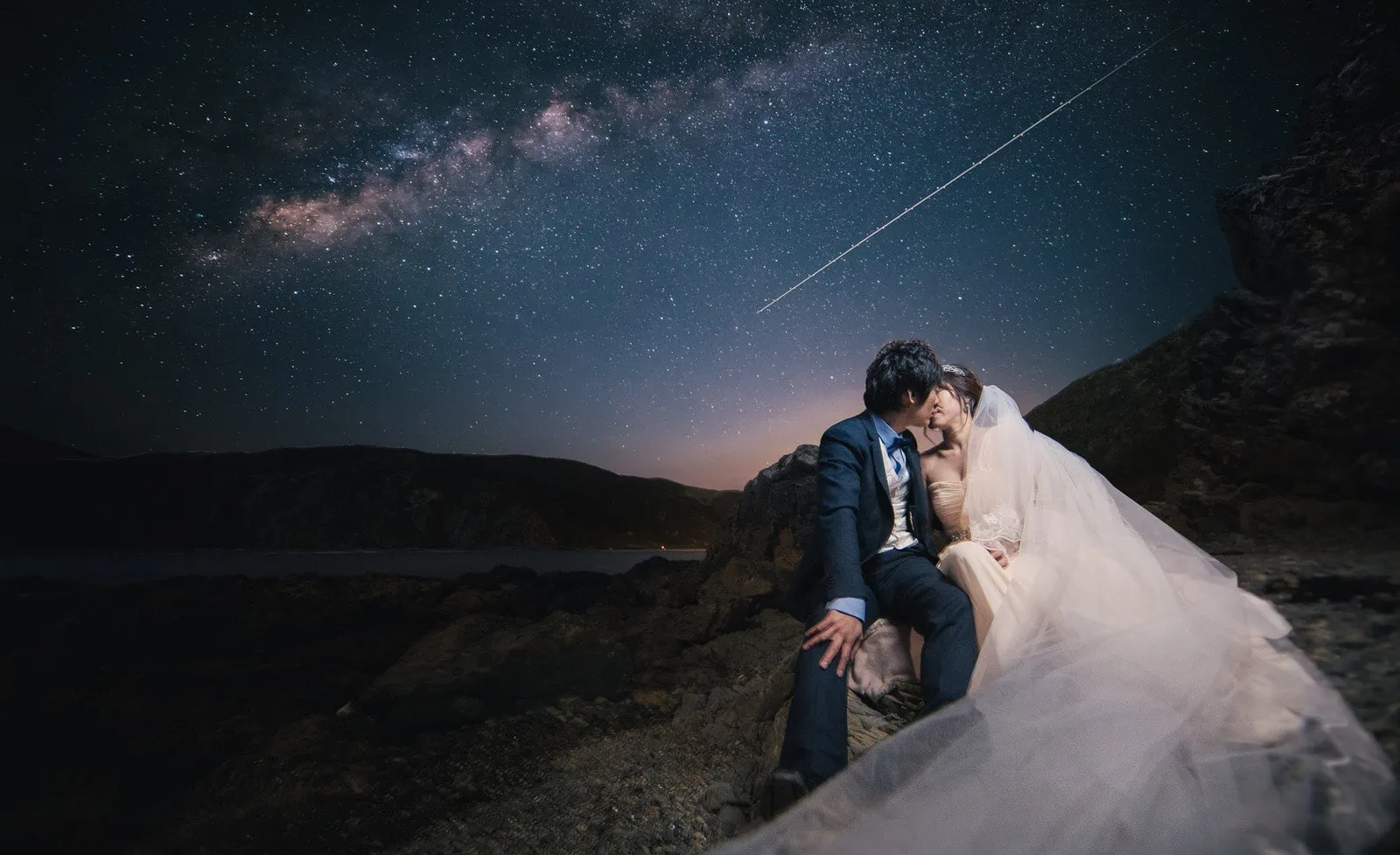 Queenstown New Zealand Elopement Wedding Photographer - A bride and groom captivated by the starry night, sitting on a rock with the milky way in the background, creating an enchanting addition to our Portfolio.