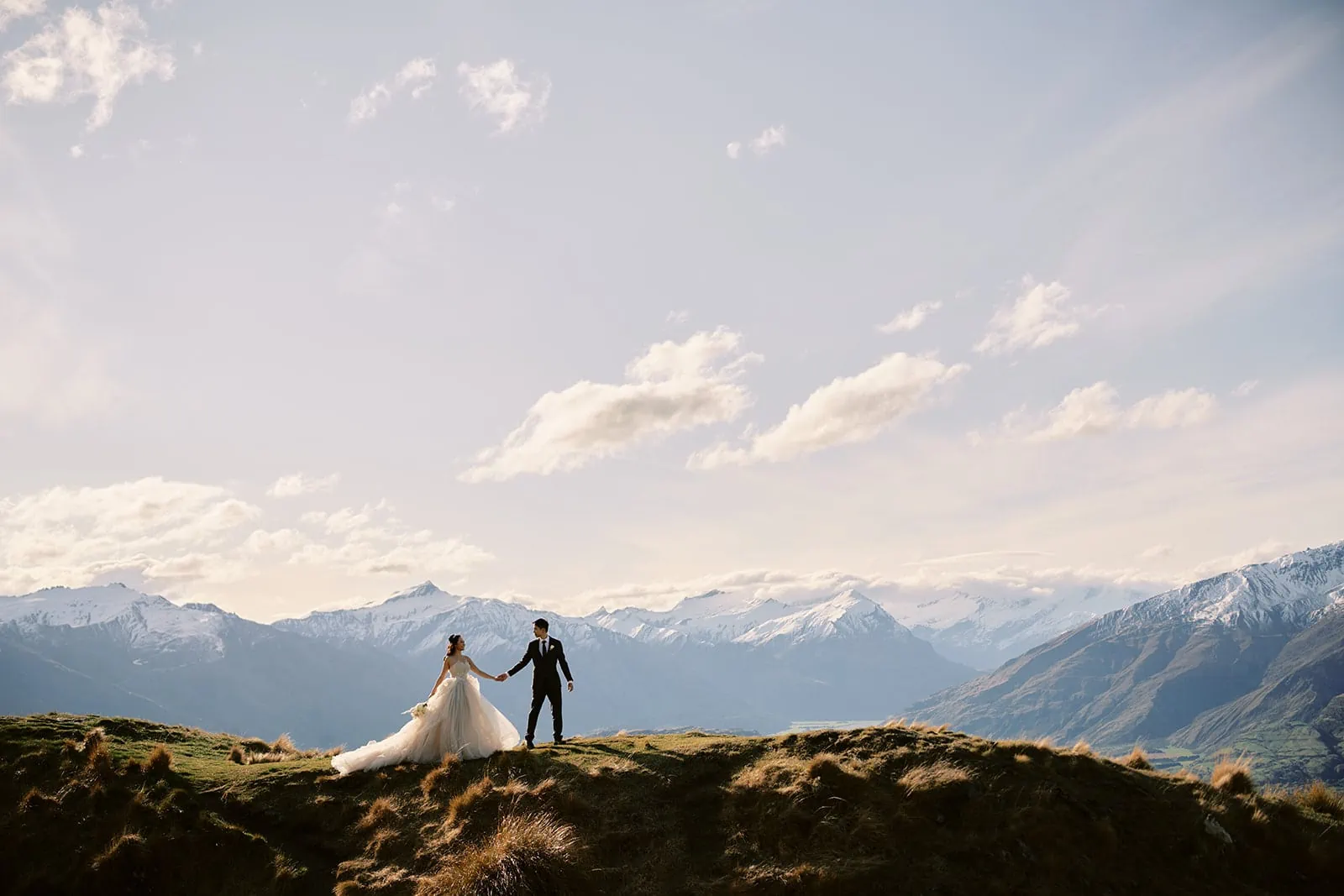 Queenstown New Zealand Elopement Wedding Photographer - A bride and groom standing on top of a hill with mountains in the background, showcasing the breathtaking scenery offered by the Queenstown heli-wedding package.