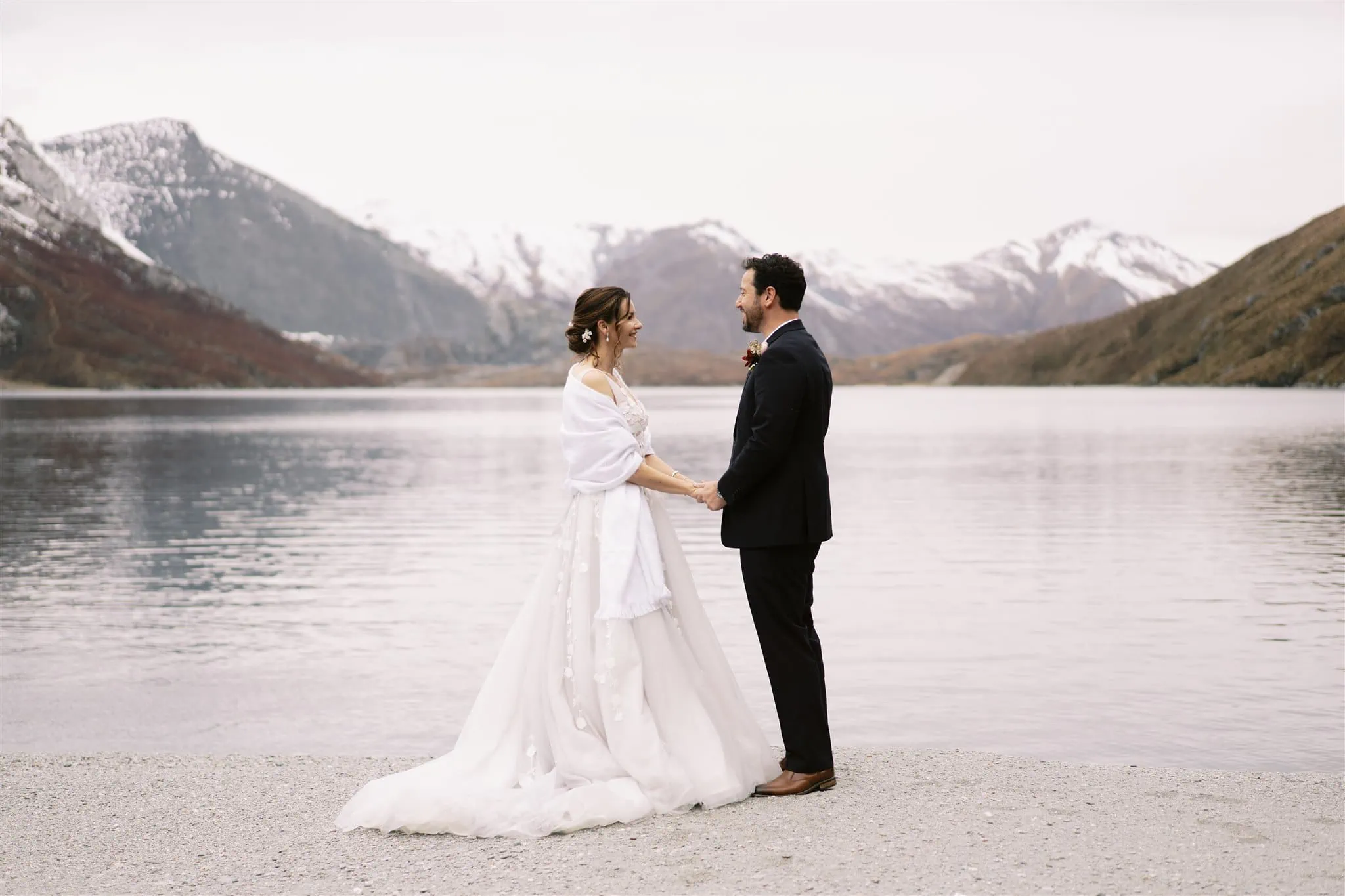 Queenstown New Zealand Elopement Wedding Photographer - Alex and Shahar, the bride and groom, standing next to a breathtaking lake with majestic mountains in the background.