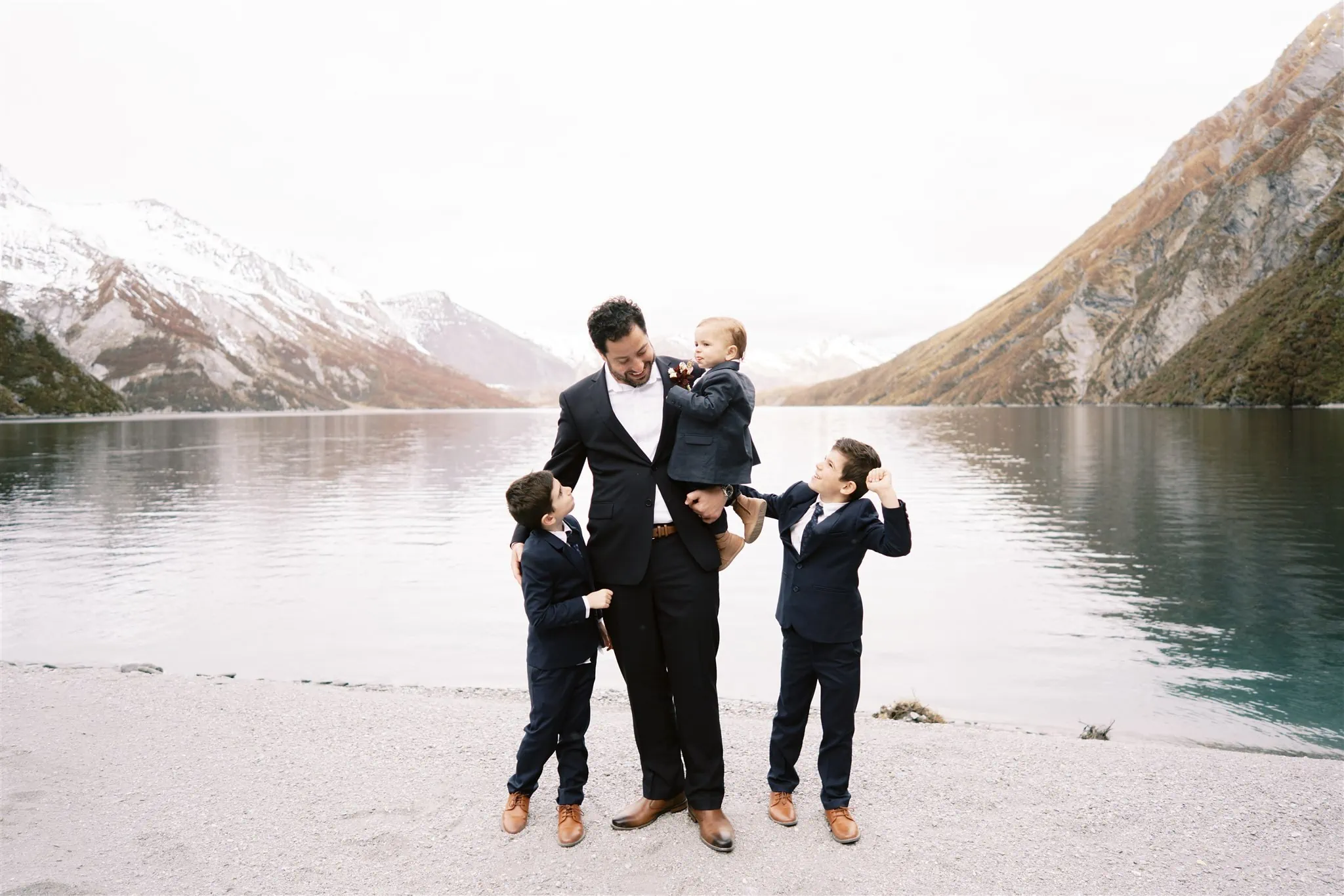 Queenstown New Zealand Elopement Wedding Photographer - Alex, a man in a suit, holds his two sons Shahar in front of a lake in New Zealand.