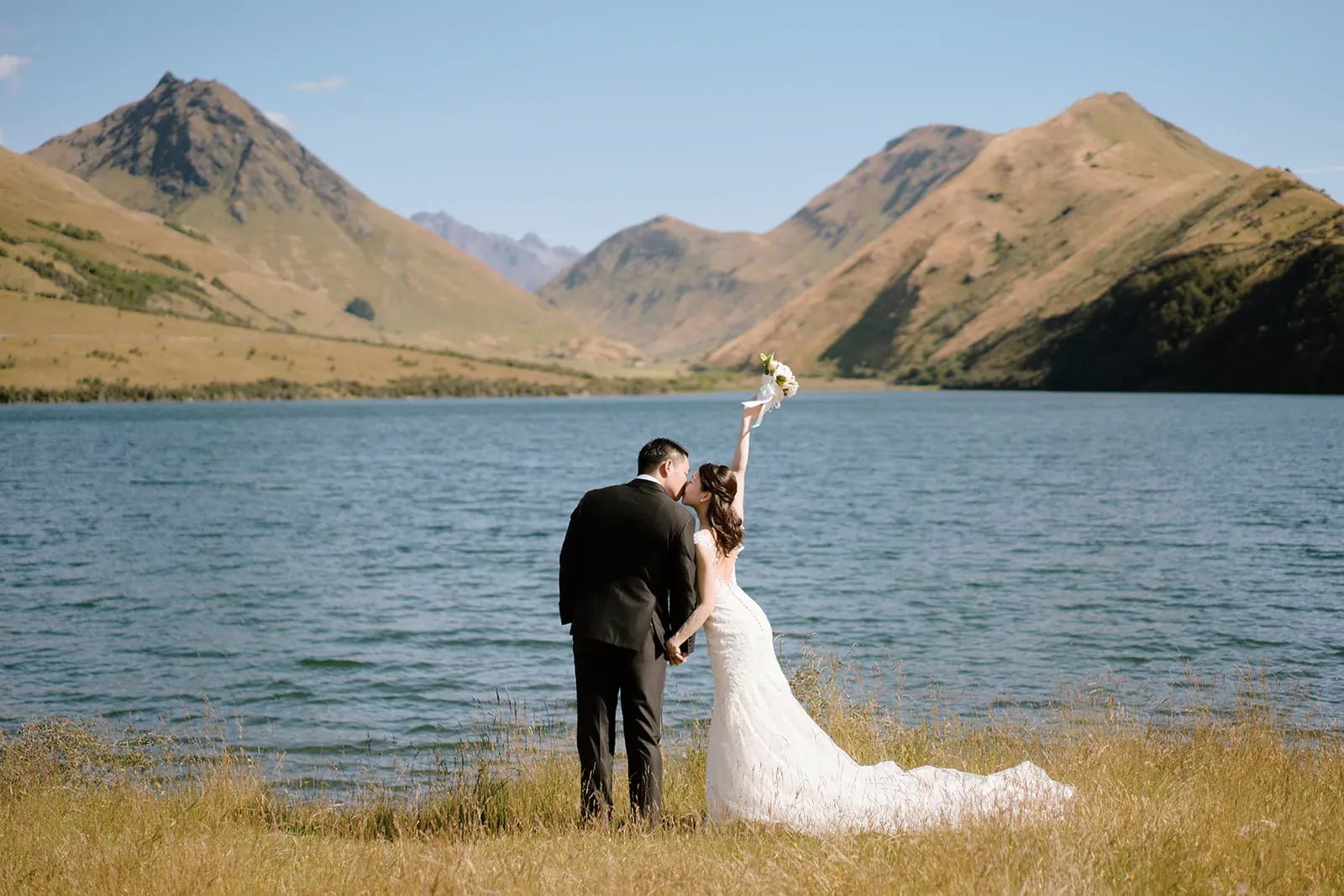 Queenstown Tekapo New Zealand Heli Wedding Elopement Pre-Wedding Shoot Photographer クイーンズタウン　テカポ　ニュージーランド　エロープメント 前撮り　フォトウェディング　結婚式 | A bride and groom standing by a lake with mountain scenery in the background for their pre-wedding shoot.