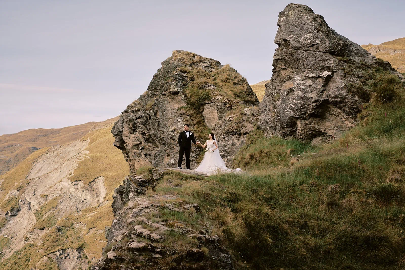 Queenstown Elopement Heli Wedding Photographer クイーンズタウン結婚式 | A bride and groom standing on the edge of a cliff in New Zealand for their pre-wedding shoot.