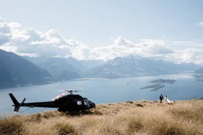 Queenstown New Zealand Elopement Wedding Photographer - A stunning bride and groom standing on a grassy hill overlooking Mt Creighton, with a helicopter nearby.