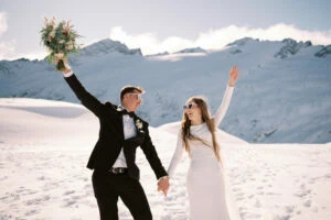 queenstown new zealand heli wedding elopement photographer videographer | A joyful bride and groom holding hands and celebrating, with the bride holding a bouquet, against a snowy Tyndall Glacier backdrop.