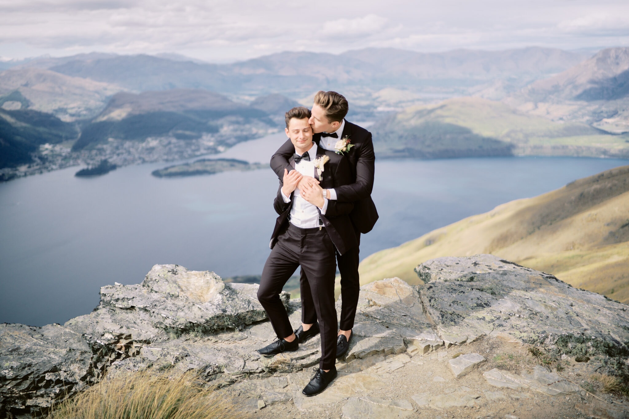 Queenstown New Zealand Elopement Wedding Photographer - Two grooms enjoying a Heli-Elopement in Queenstown, hugging on top of a mountain overlooking lake wanaka.