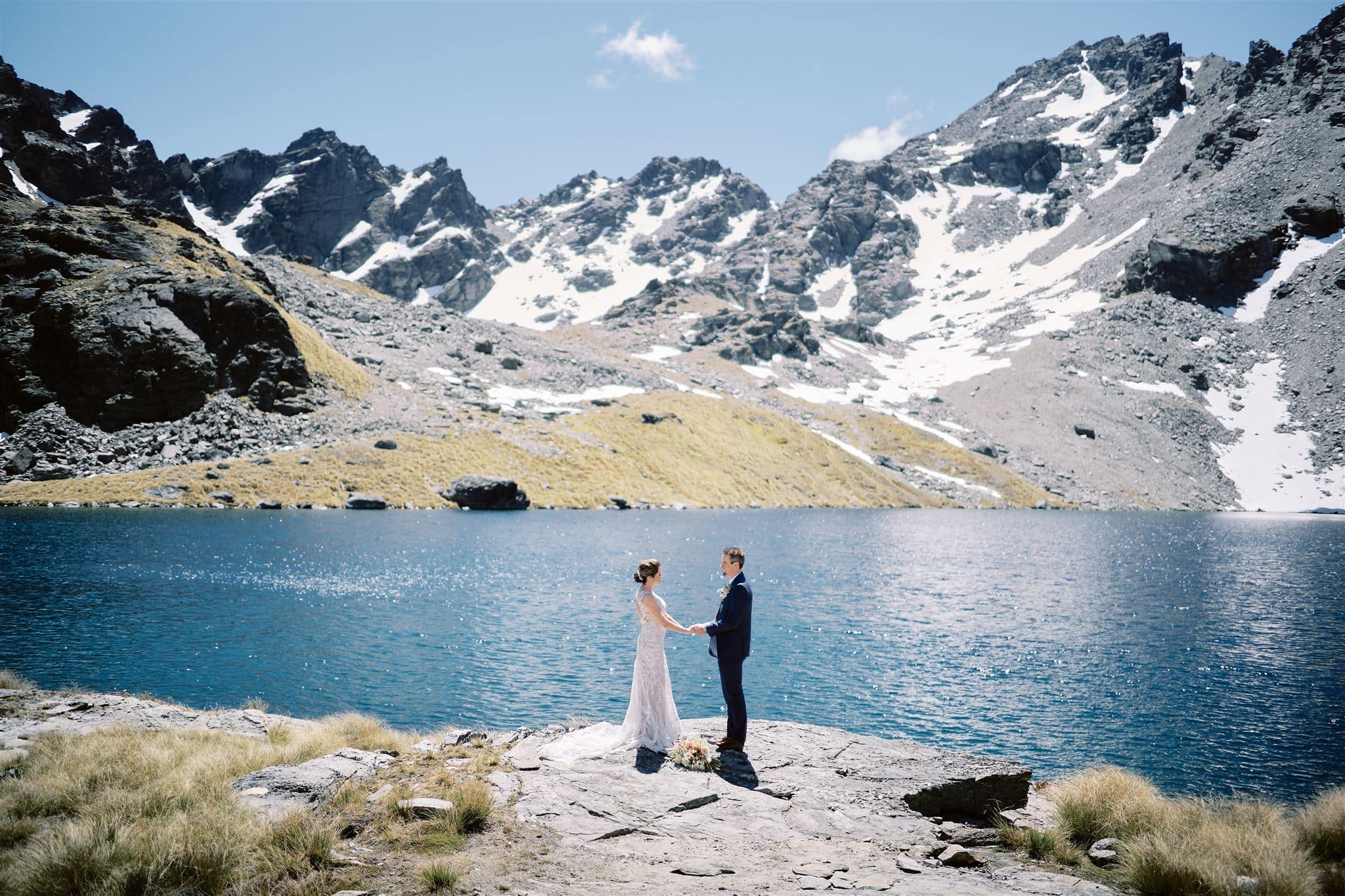 Queenstown New Zealand Elopement Wedding Photographer - A bride and groom happily stand next to a serene lake in the beautiful mountains of Queenstown, one of the top heli-wedding locations and destinations.