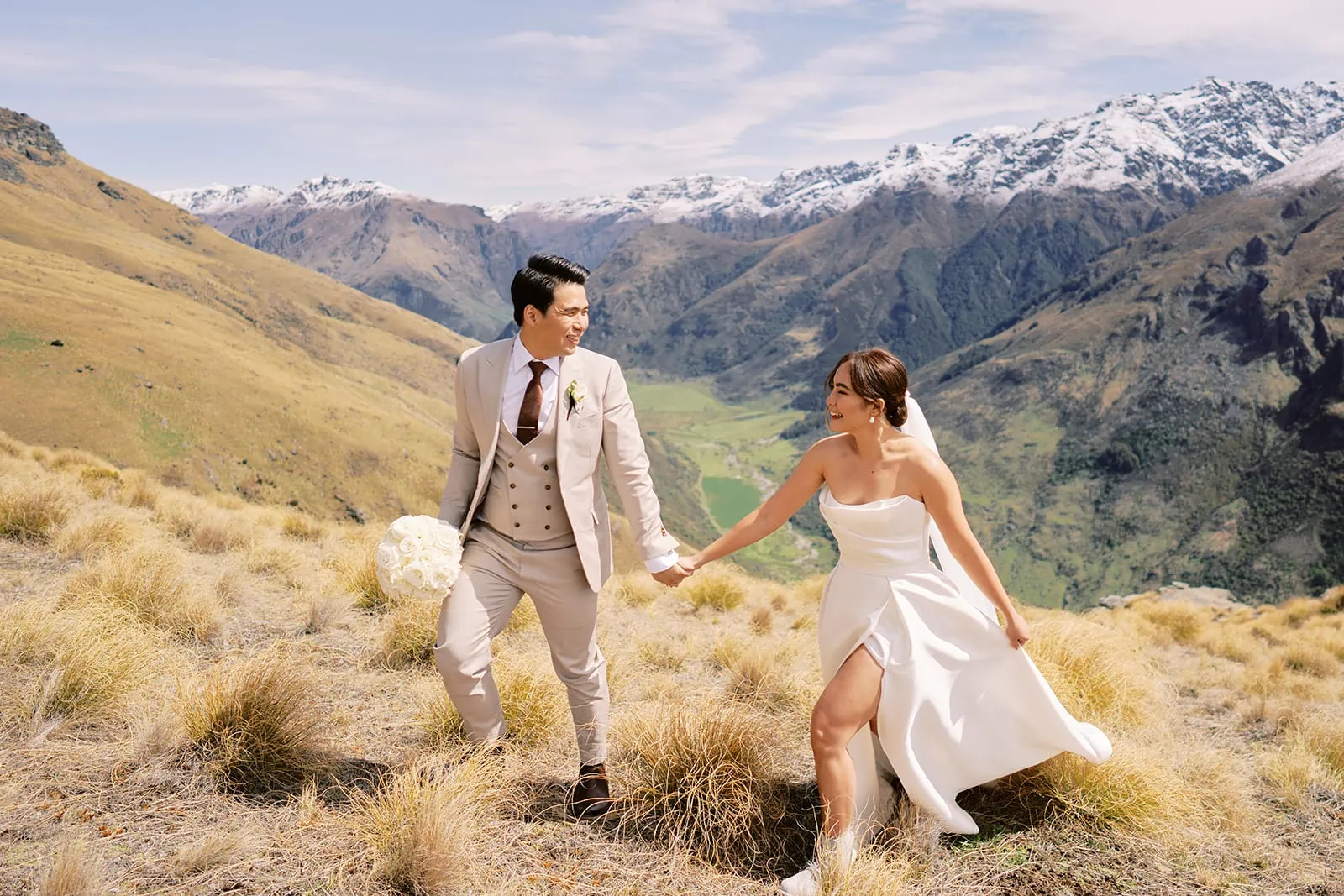 Queenstown Elopement Heli Wedding Photographer クイーンズタウン結婚式 | A Queenstown wedding couple, Tobi and Ceidi, strolling through a picturesque grassy field with majestic mountains as their backdrop.