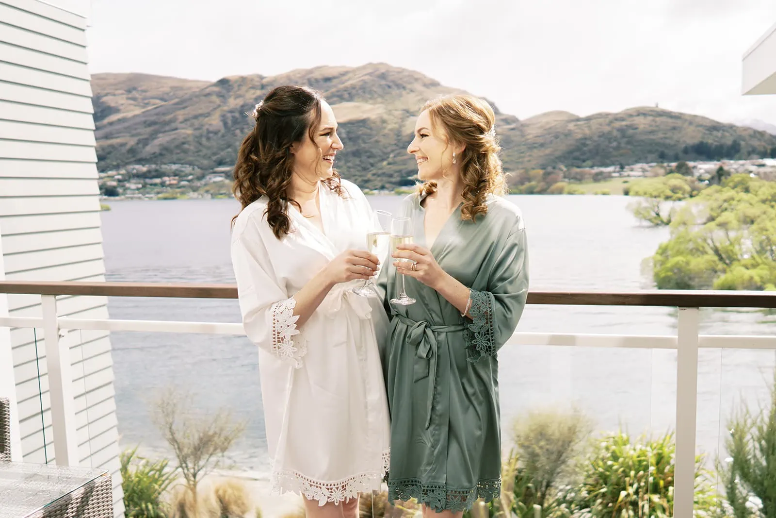 Queenstown Wedding Photographer Nat & Tim, a couple celebrating their Queenstown EloPement Wedding, are seen on a balcony overlooking Lake Wanaka. They are happily sipping champagne while wearing robes, enjoying the picturesque