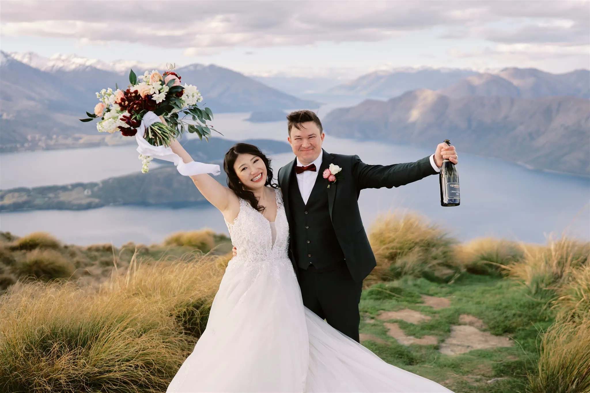 Queenstown Elopement Heli Wedding Photographer クイーンズタウン結婚式 | An elopement wedding on top of a mountain in New Zealand, with the bride and groom holding a bottle of champagne to celebrate.