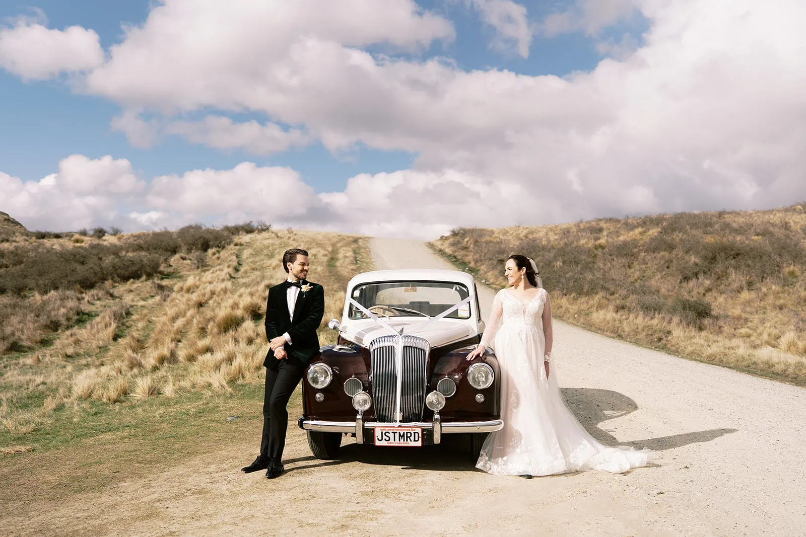 Queenstown Wedding Photographer Nat & Tim, a bride and groom, standing next to a vintage car on a dirt road during their Queenstown elopement wedding at Deer Park Heights.