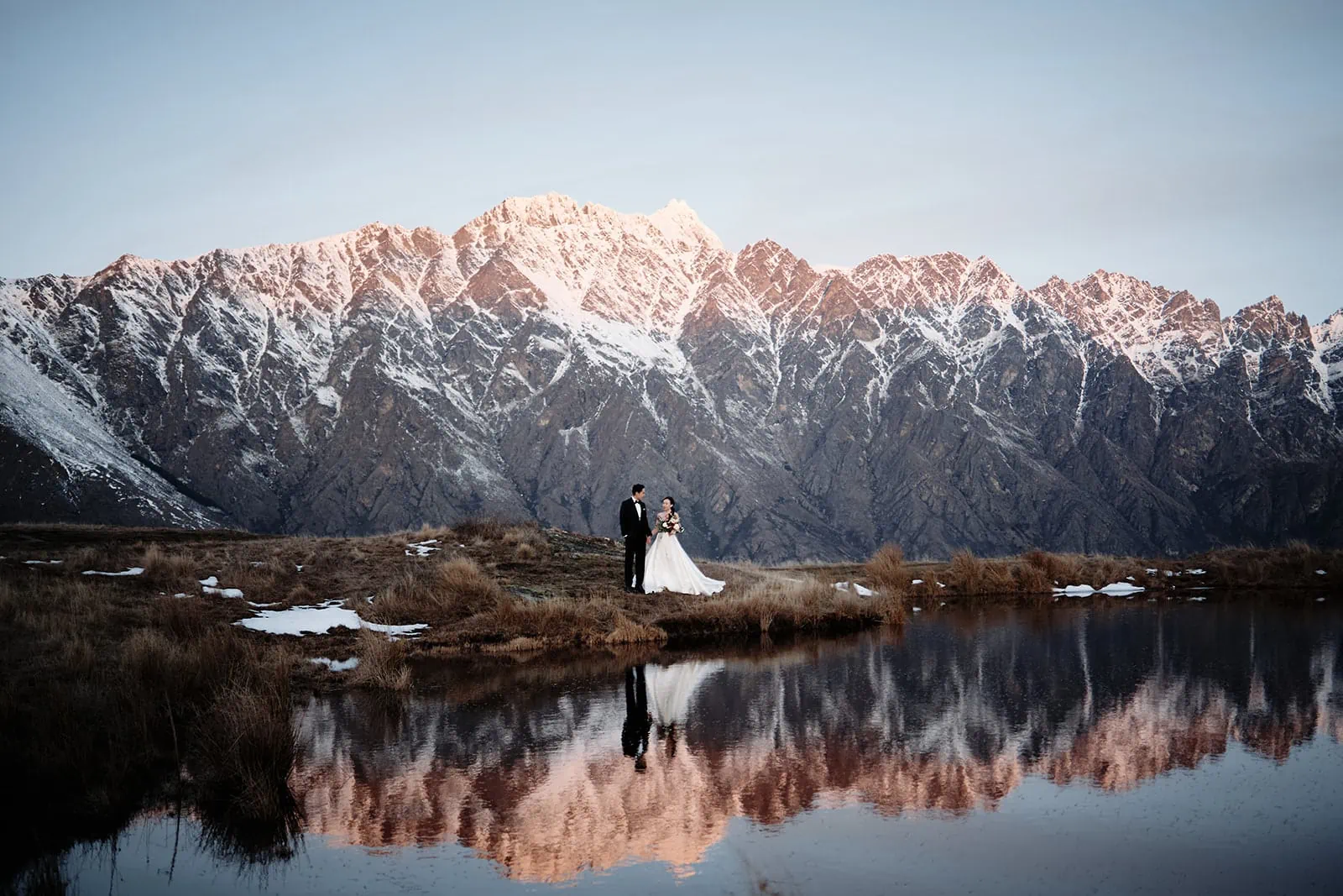 Queenstown Wedding Photographer A bride and groom enjoying a queenstown heli-wedding package, standing in front of a lake with mountains in the background.