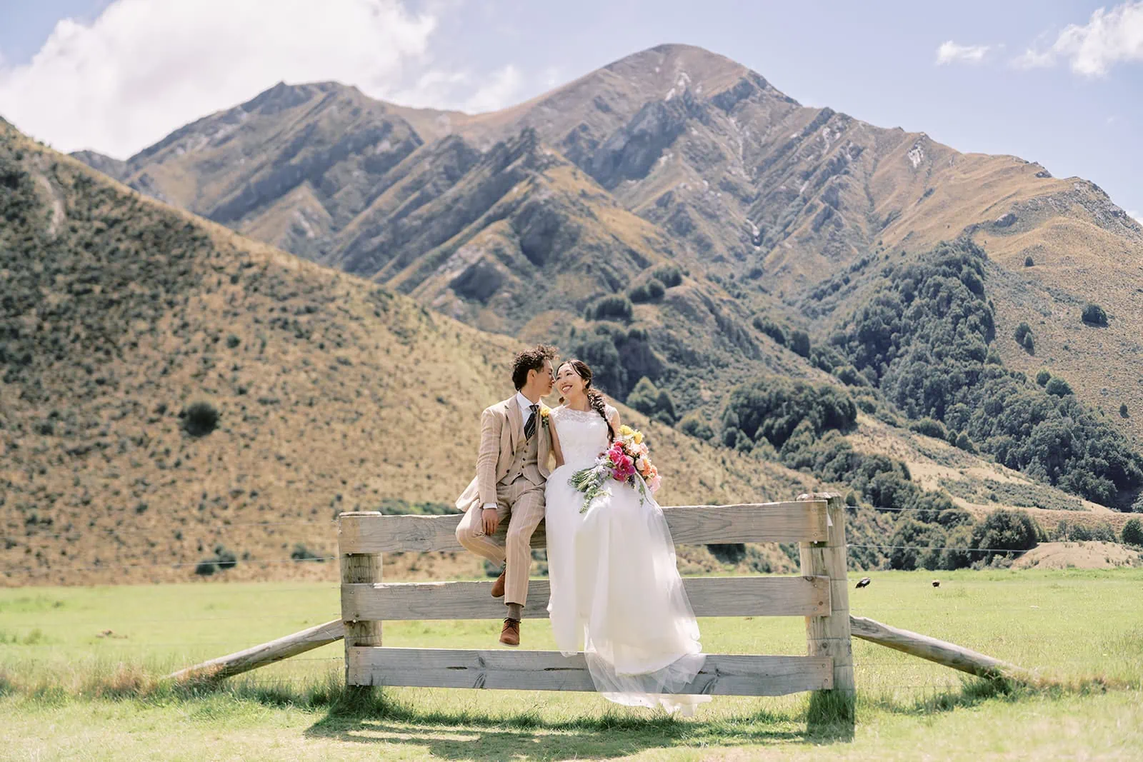Queenstown Elopement Heli Wedding Photographer クイーンズタウン結婚式 | Saki & Taisei enjoying a Queenstown Heli Pre-Wedding Photoshoot on a fence in a field with mountains in the background.