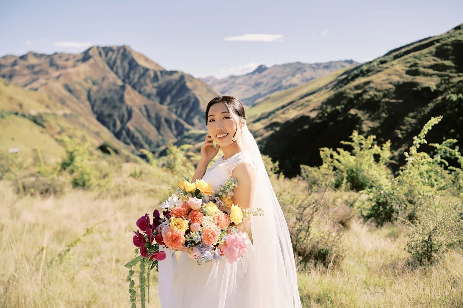 Queenstown Elopement Heli Wedding Photographer クイーンズタウン結婚式 | Saki & Taisei having a pre-wedding photoshoot in Queenstown Heli, with the bride holding a bouquet in a field and mountains in the background.