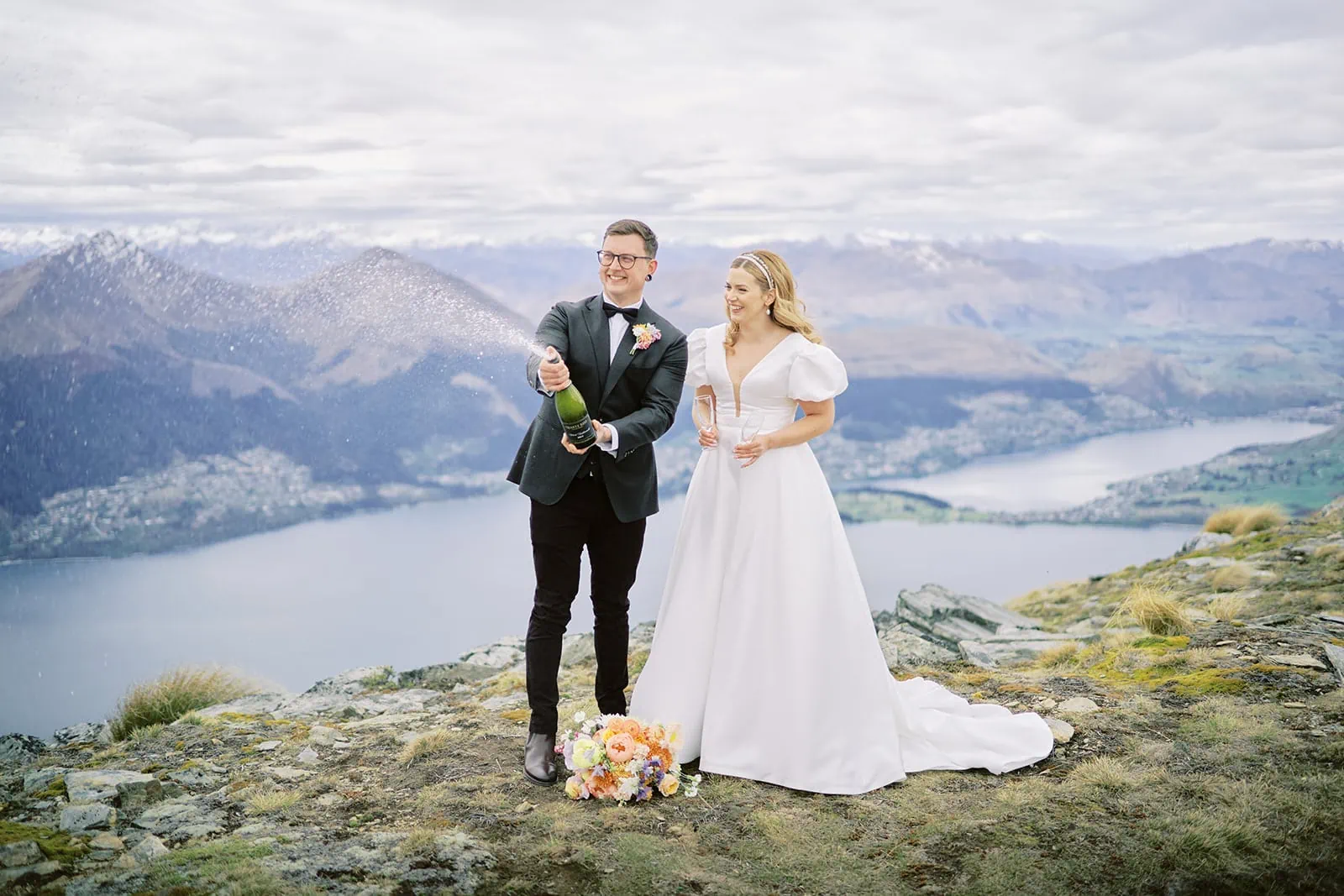 Queenstown Elopement Heli Wedding Photographer クイーンズタウン結婚式 | Sophie and Thomas, a bride and groom, standing on top of Queenstown's majestic mountain with a bottle of champagne.