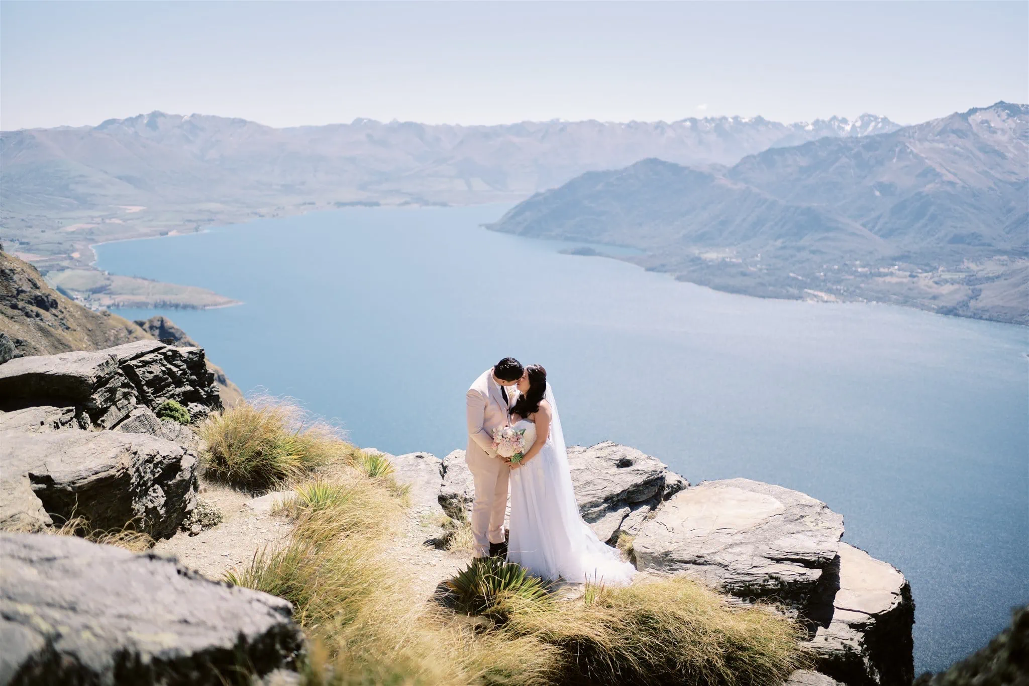 Queenstown Elopement Heli Wedding Photographer クイーンズタウン結婚式 | A Queenstown elopement of a man and woman kissing on a cliff overlooking a lake.