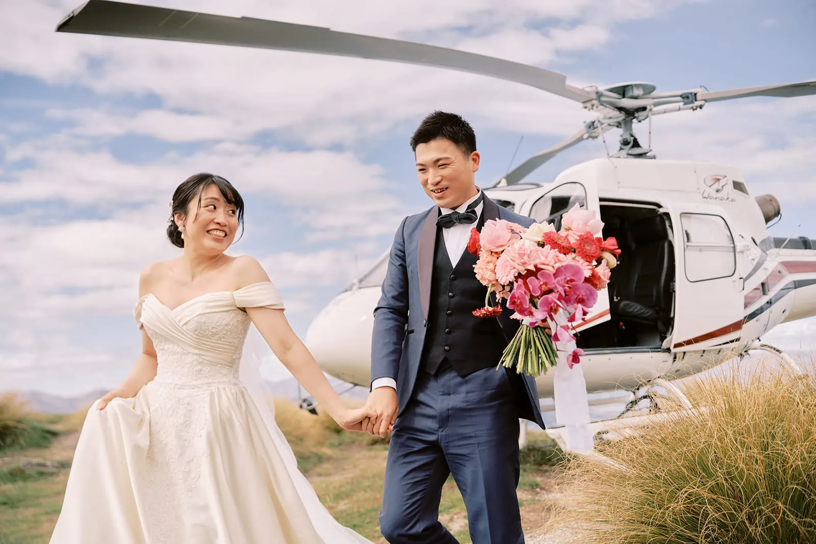 Queenstown Elopement Heli Wedding Photographer クイーンズタウン結婚式 | A bride and groom partaking in a pre-wedding photoshoot, holding hands in front of a helicopter.