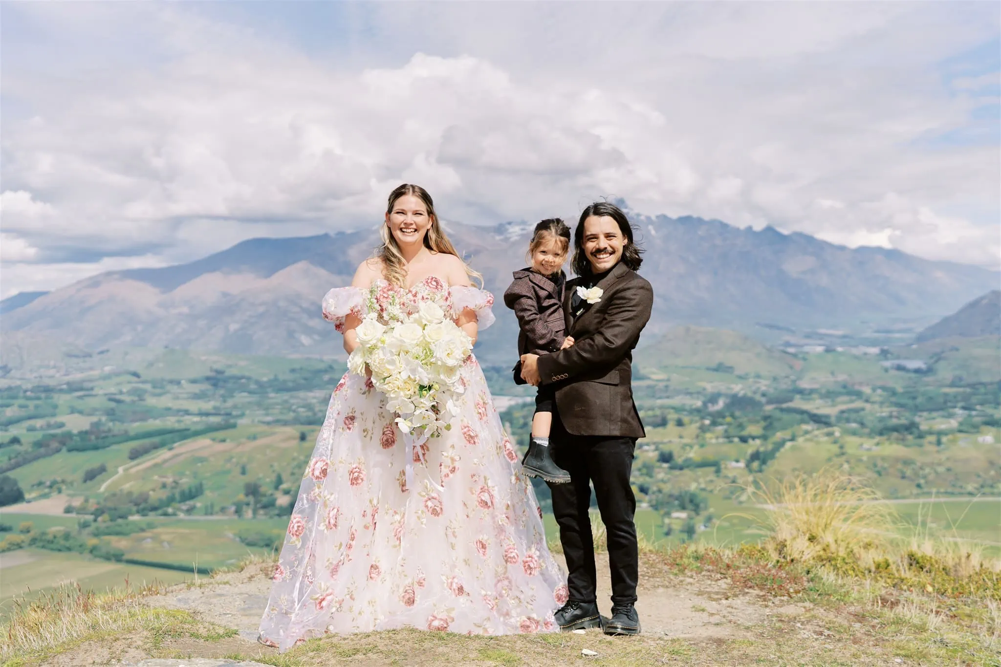 Queenstown Elopement Heli Wedding Photographer クイーンズタウン結婚式 | A Coronet Peak elopement with a bride and groom standing on top of a mountain, accompanied by a child.